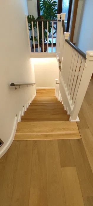Widok z góry na drewniane schody po renowacji, z jasnymi stopniami i białymi balustradami, prowadzące w dół, z widocznym fragmentem drewnianej podłogi na pierwszym planie i rośliną doniczkową...
