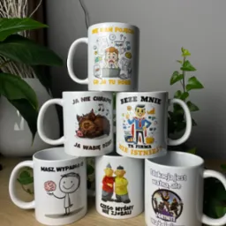 Piramida z sześciu białych kubków z różnymi nadrukami, w tym motywami humorystycznymi, zwierzęcymi i nawiązującymi do gier komputerowych, ustawiona na drewnianym blacie na tle roślin doniczkowych...