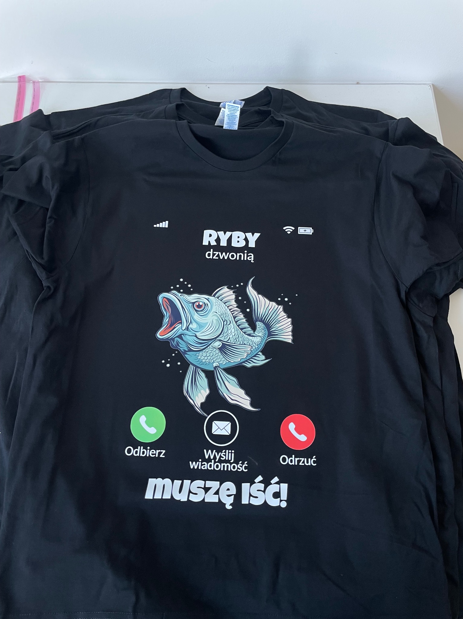 Stos czarnych koszulek z nadrukiem imitującym ekran smartfona z dzwoniącą rybą i tekstem 'RYBY dzwonią muszę iść!'