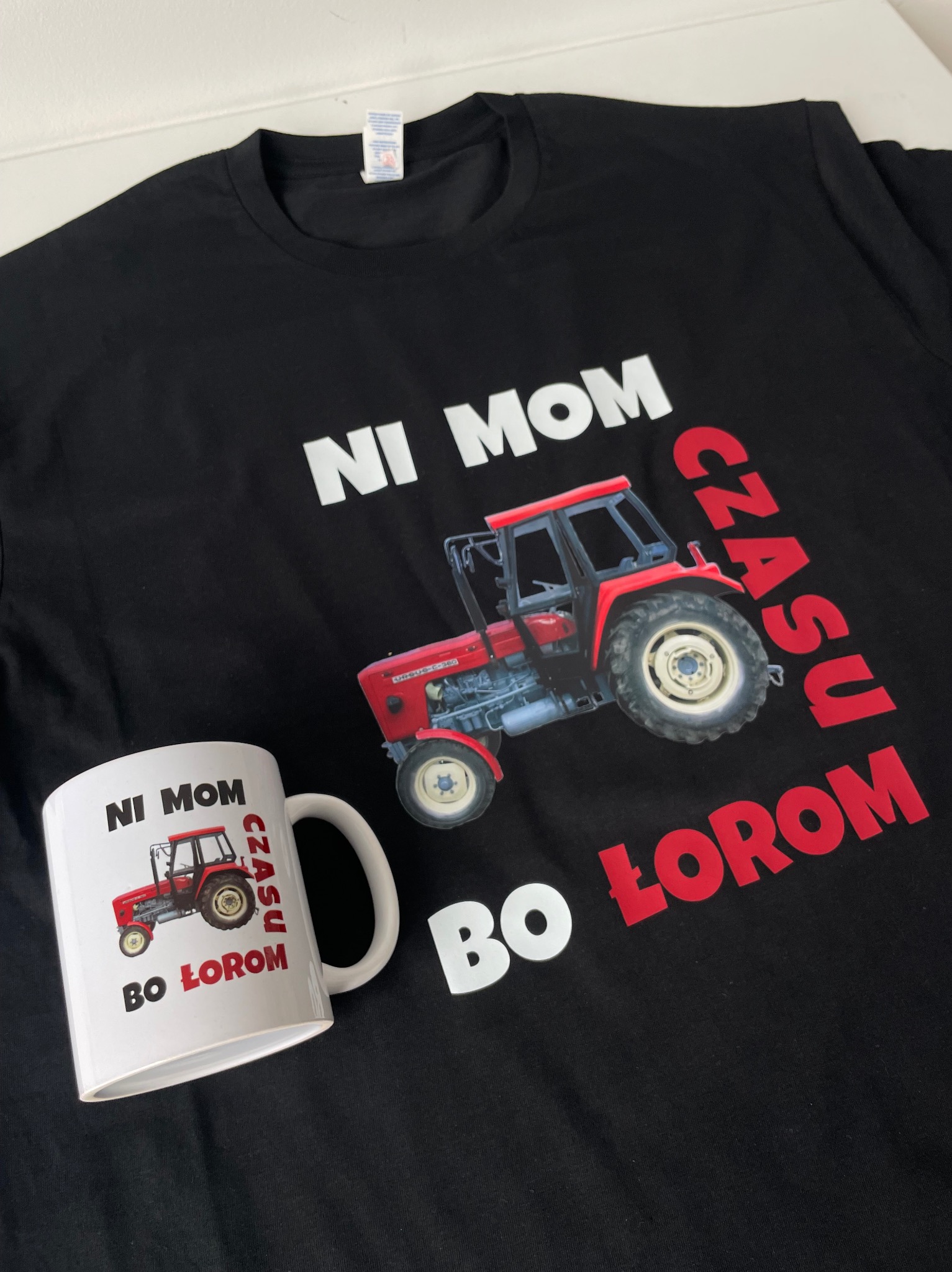 Czarna koszulka i biały kubek z nadrukiem czerwonego traktora Ursus C-360 i napisem 'NI MOM CZASU BO ŁOROM' leżące na białym tle.