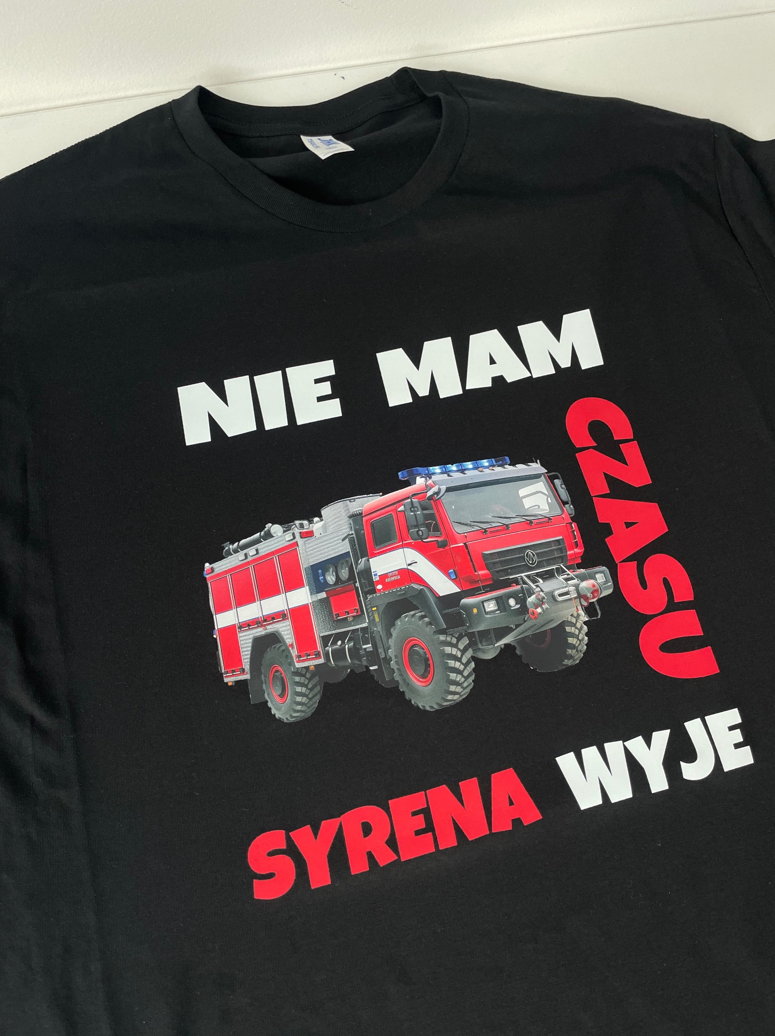 Czarna koszulka z nadrukiem wozu strażackiego, napisem 'NIE MAM CZASU SYRENA WYJE' w kolorach białym i czerwonym, leżąca na białym tle.