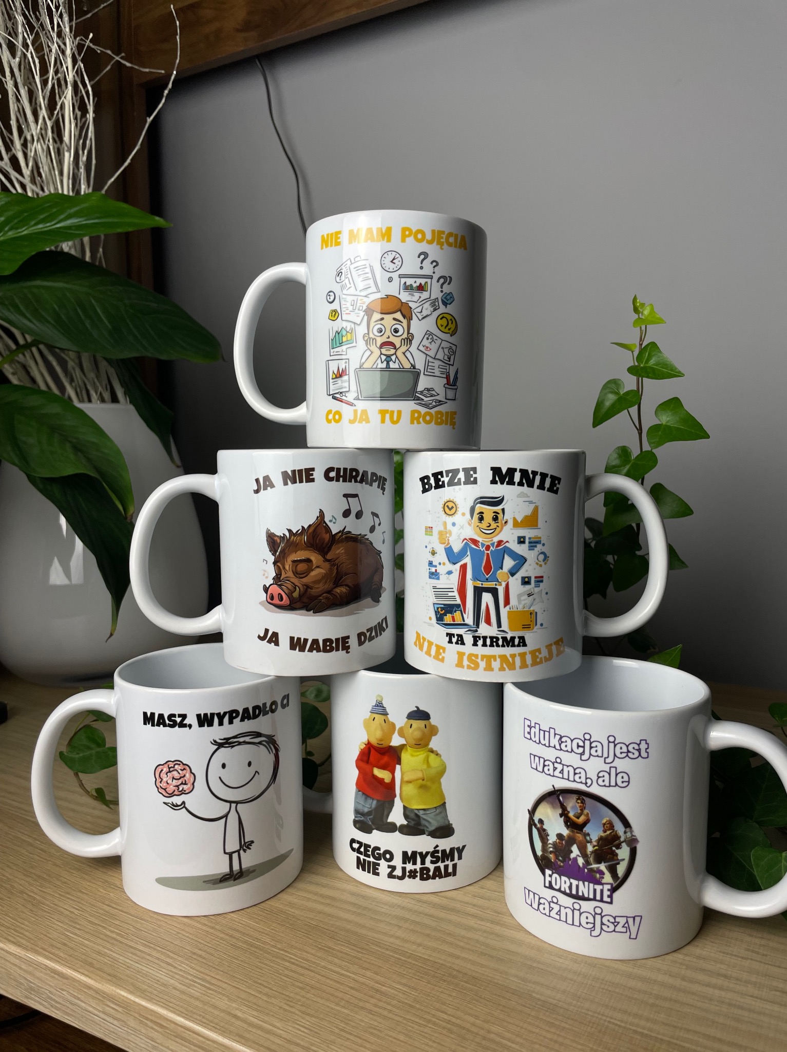 Piramida z sześciu białych kubków z różnymi nadrukami, w tym motywami humorystycznymi, zwierzęcymi i nawiązującymi do gier komputerowych, ustawiona na drewnianym blacie na tle roślin doniczkowych...