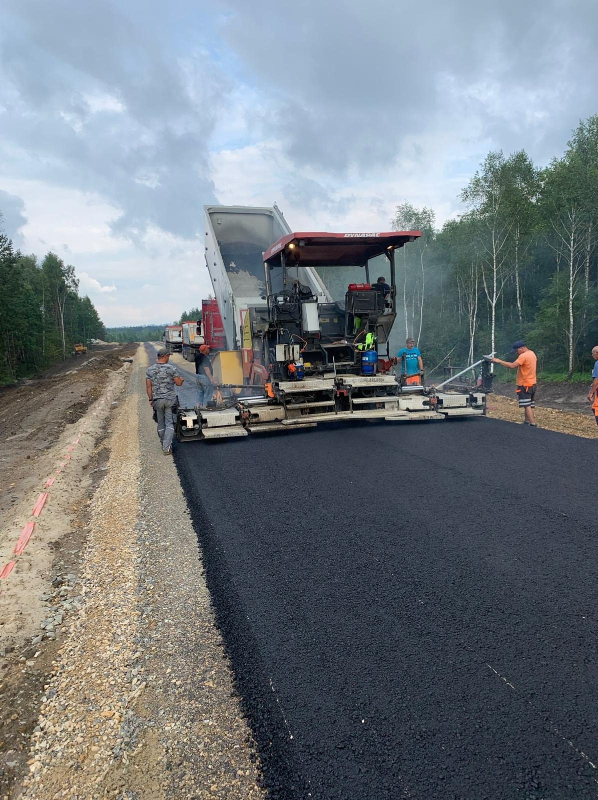 Układanie asfaltu na drodze w Moskorzewie. Widoczny finiszer do asfaltu Dynapac i pracownicy w odblaskowych ubraniach. W tle drzewa i ciężarówki.