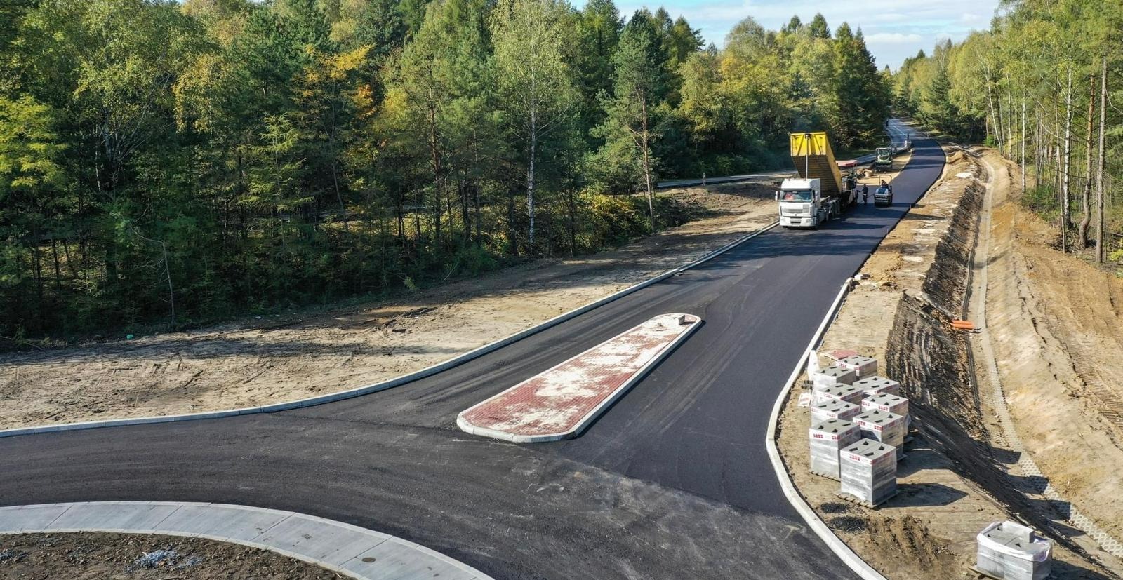 Nowa asfaltowa droga z wysepką dla pieszych i paletami kostki brukowej na poboczu, w tle ciężarówka z wywrotką i maszyny budowlane, otoczenie leśne.