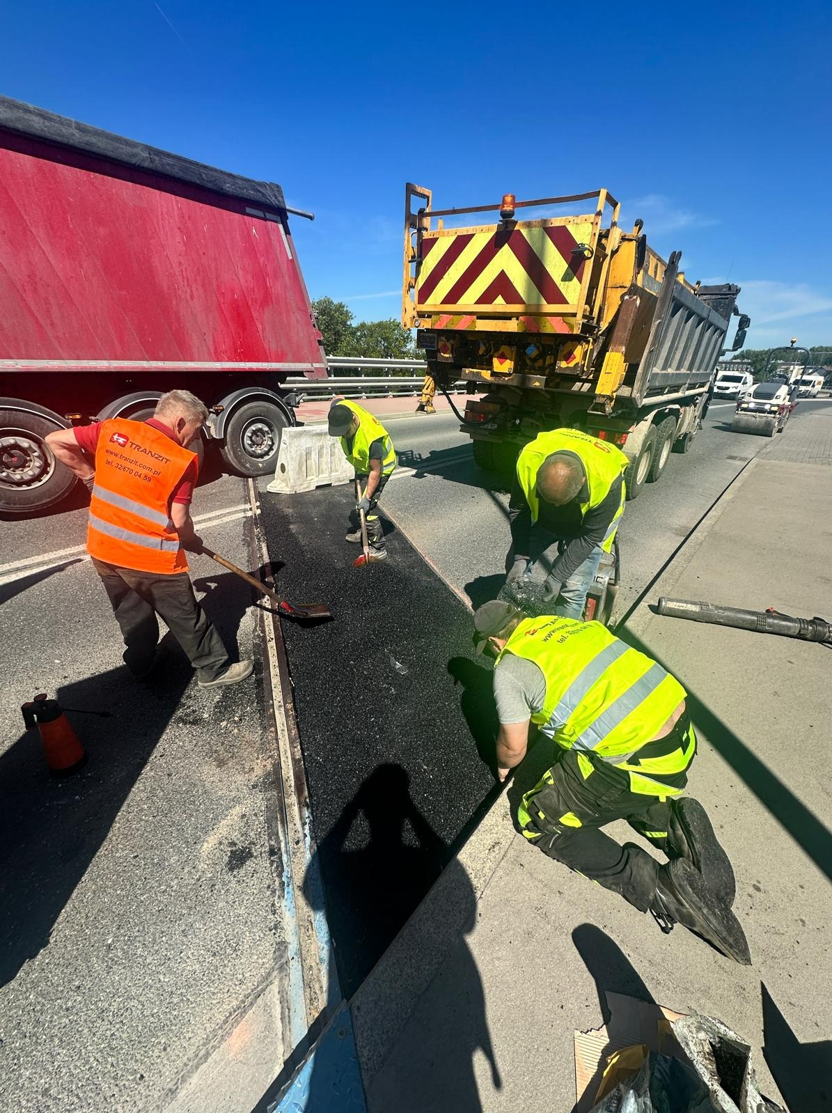 Robotnicy w jaskrawych kamizelkach układają asfalt na drodze, widoczny czerwony samochód ciężarowy i żółta wywrotka z migającym światłem ostrzegawczym, w tle ruch uliczny.