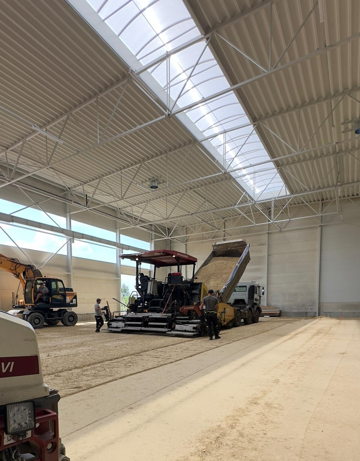 Wnętrze hali z maszynami do utwardzania podłoża: rozściełacz asfaltu Dynapac, wywrotka z piaskiem i koparka Hyundai. Dwóch pracowników nadzoruje proces.