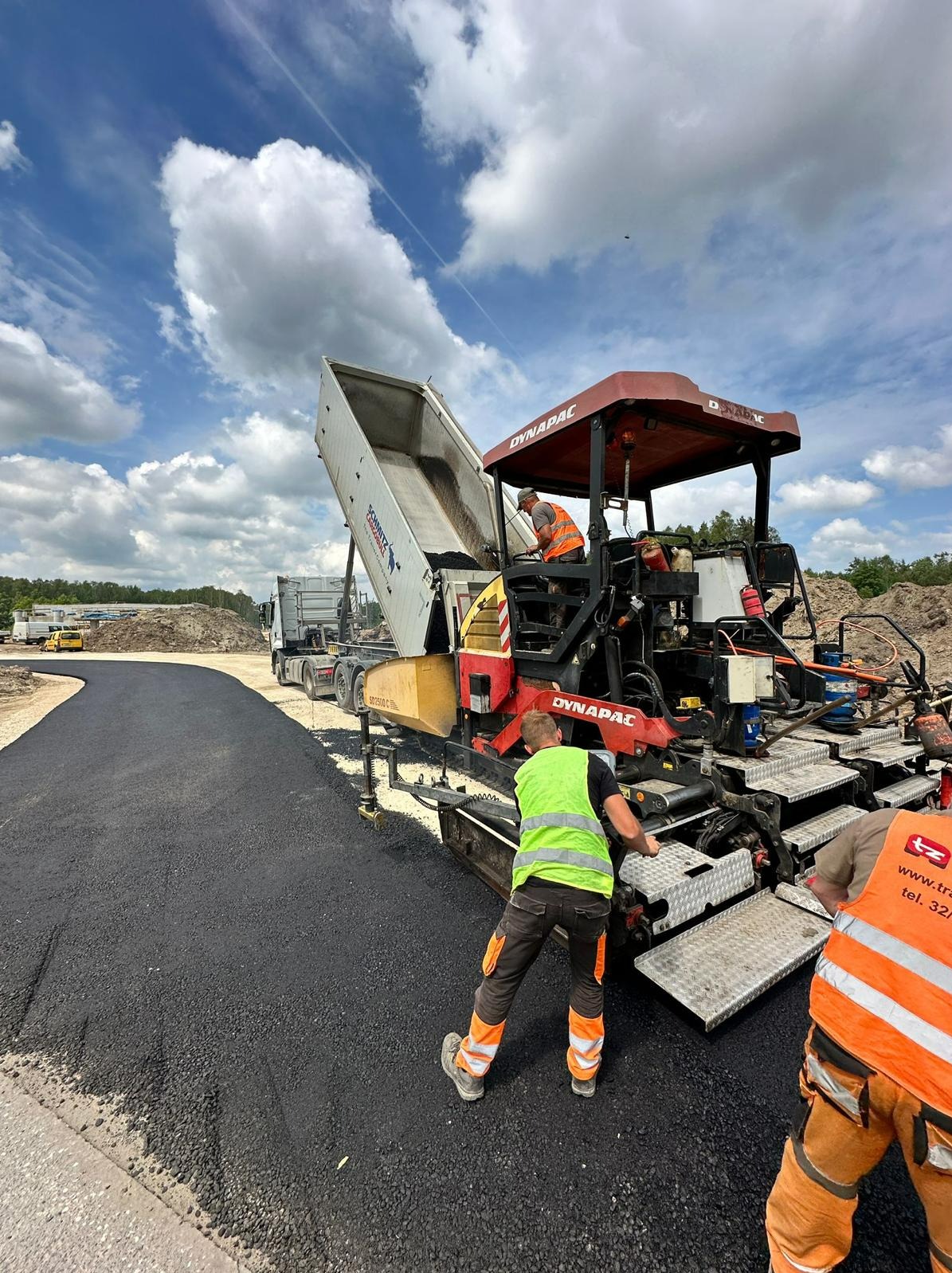 Układanie asfaltu na drodze przy użyciu maszyny Dynapac, z widocznym pracownikiem w kamizelce odblaskowej i wywrotką z materiałem, na tle błękitnego nieba z chmurami.