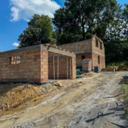 Surowy stan budynku z czerwonej cegły w trakcie budowy, widoczne drewniane podpory i palety z materiałami budowlanymi na nier&oacute;wnym, błotnistym terenie w słoneczny dzień.