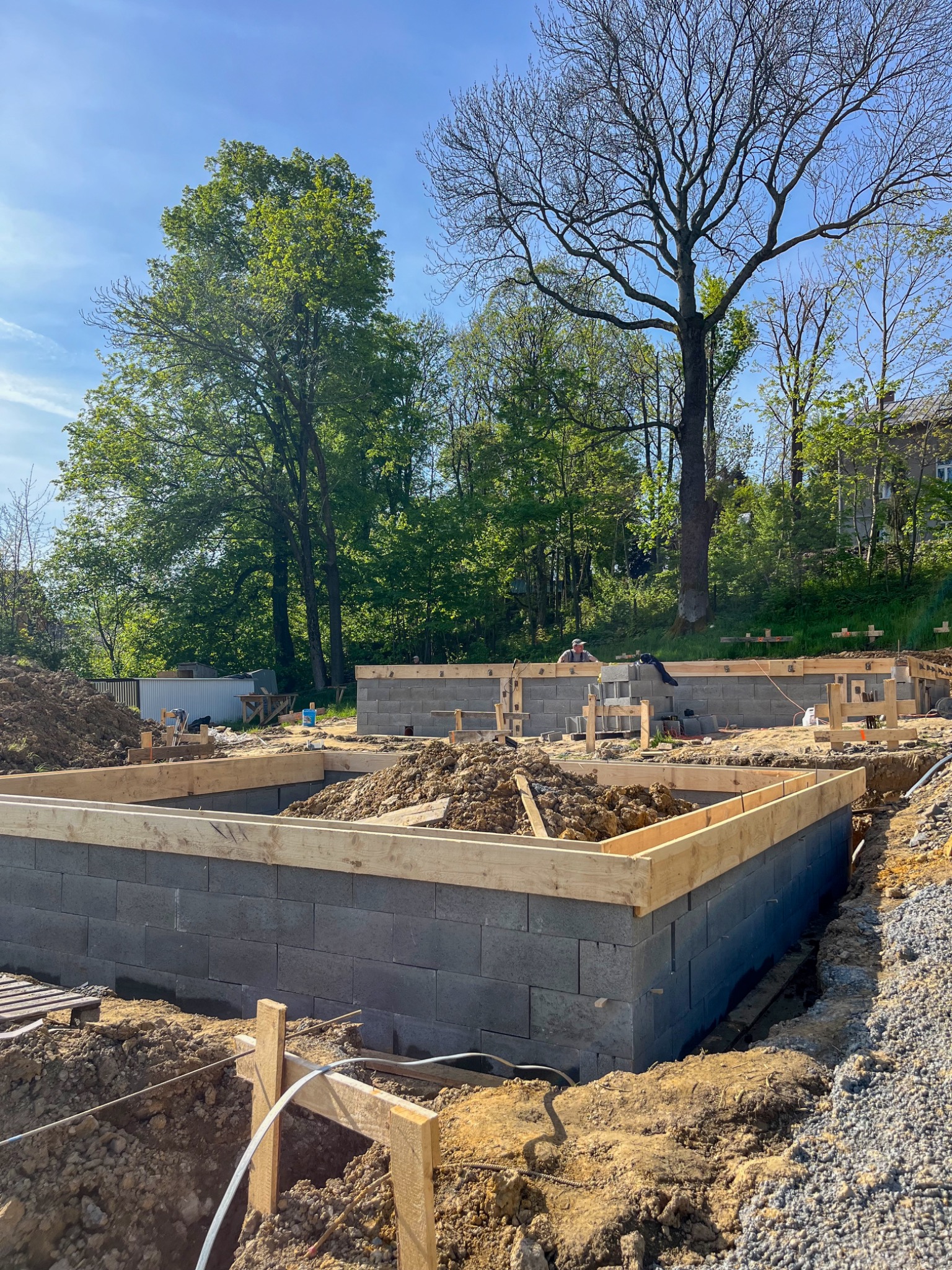 Wykop pod fundamenty z wzniesionymi ścianami z betonowych bloczków, drewnianym szalunkiem i pracującym w tle mężczyzną, w otoczeniu drzew i błękitnego nieba.