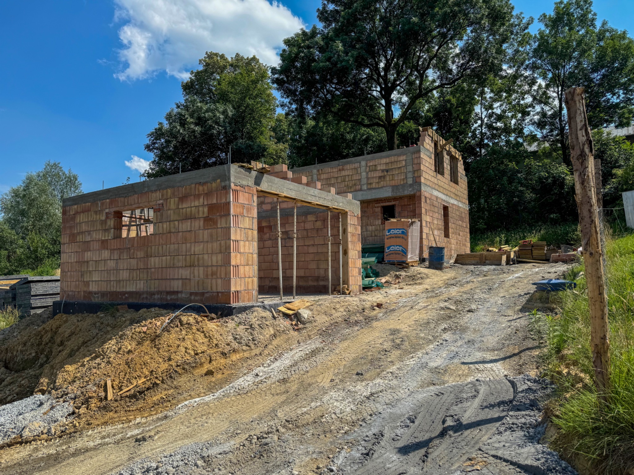 Surowy stan budynku z czerwonej cegły w trakcie budowy, widoczne drewniane podpory i palety z materiałami budowlanymi na nierównym, błotnistym terenie w słoneczny dzień.