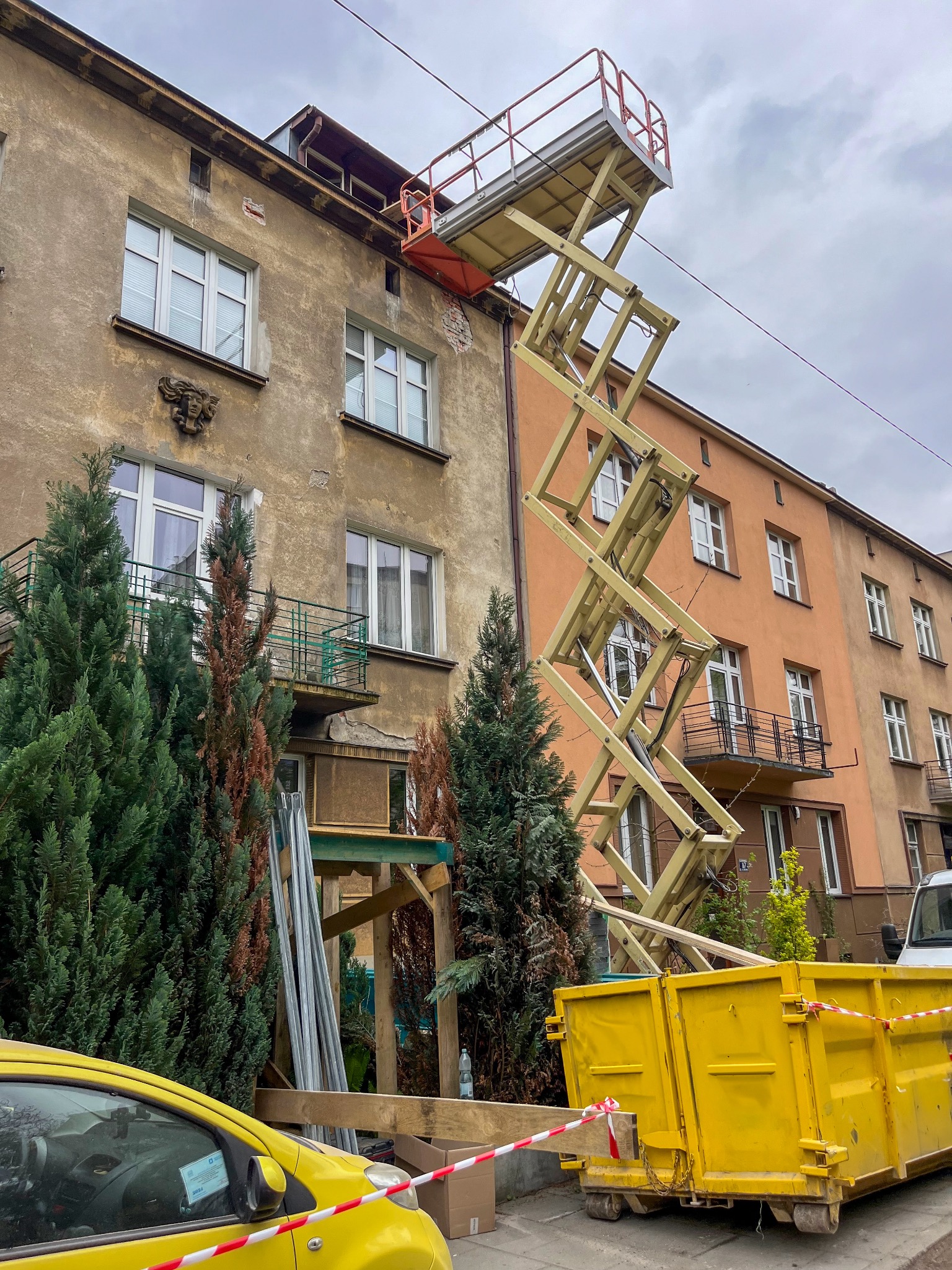 Remont elewacji kamienicy z użyciem podnośnika nożycowego i kontenera na gruz, widoczne rusztowania i materiały budowlane.