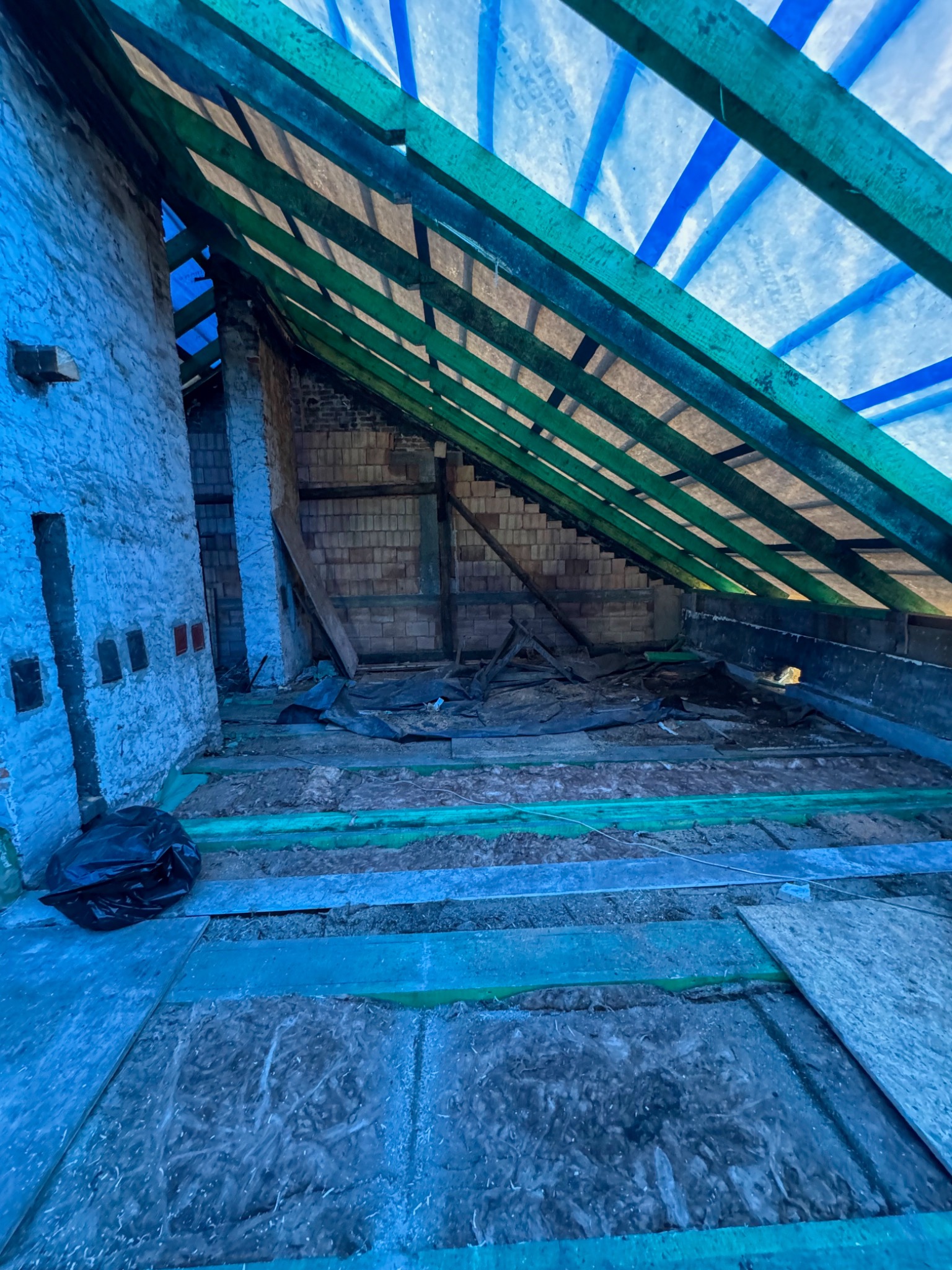 Wnętrze poddasza w trakcie remontu: widoczne krokwie z folią, częściowo ułożona izolacja podłogowa, fragment murowanej ściany z otworami okiennymi i czarny worek na śmieci w rogu.