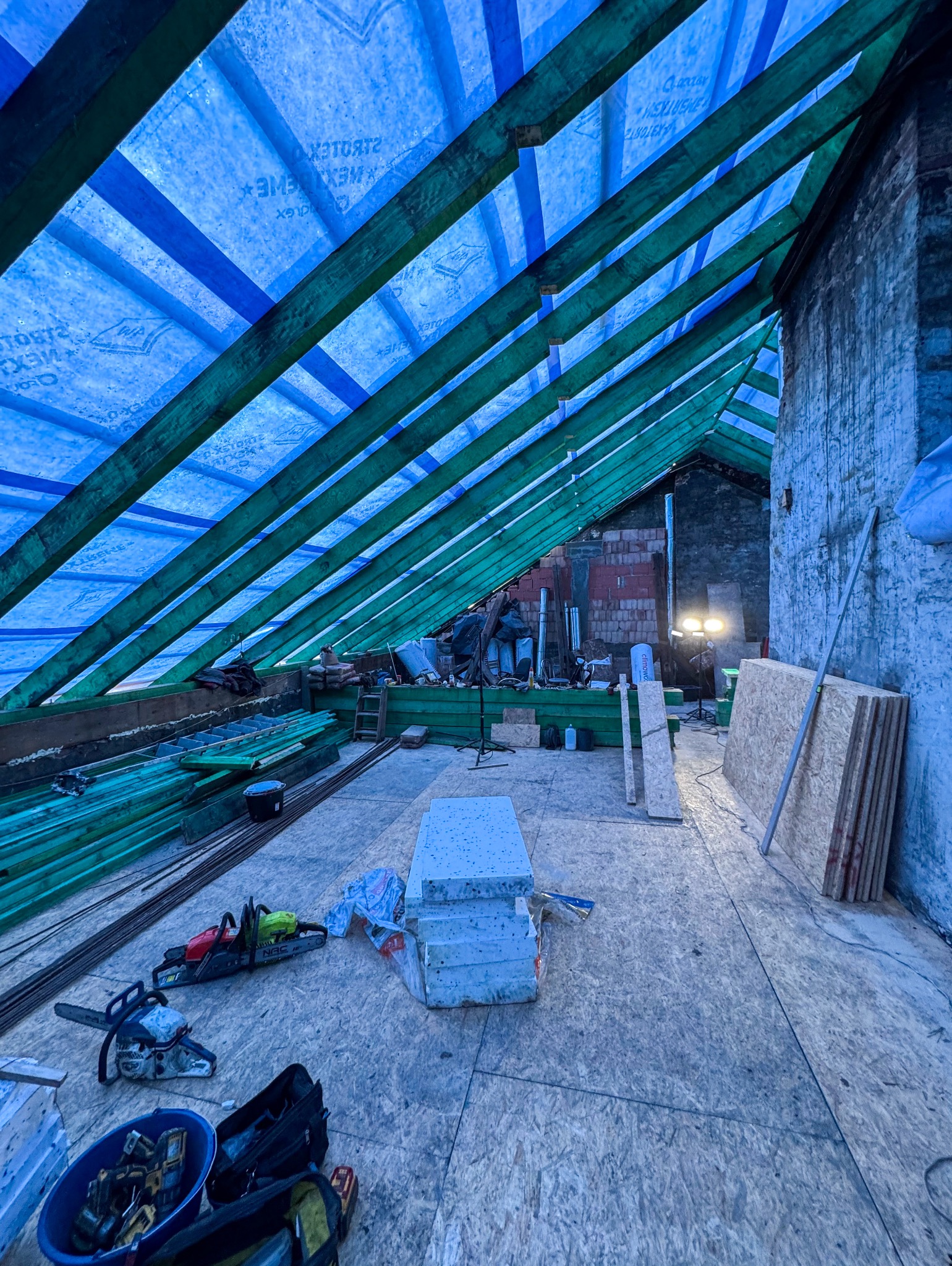 Wnętrze poddasza w trakcie remontu z widoczną konstrukcją dachu zabezpieczoną niebieską folią, narzędzia budowlane, deski i materiały izolacyjne na drewnianej podłodze.