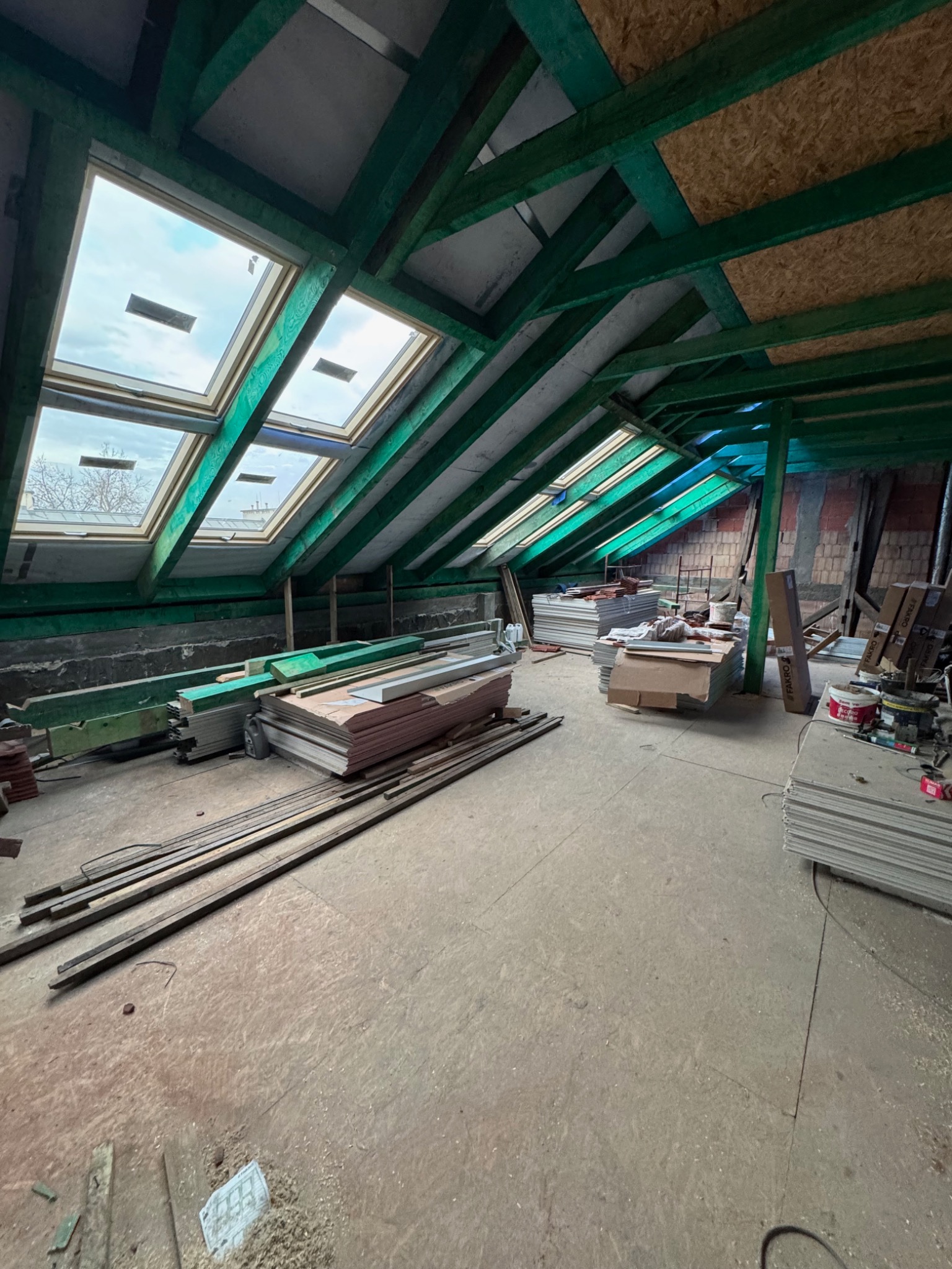 Poddasze w trakcie adaptacji w Nowym Sączu, widoczne odeskowanie dachu z oknami połaciowymi, zielone belki konstrukcyjne i materiały budowlane składowane na podłodze.