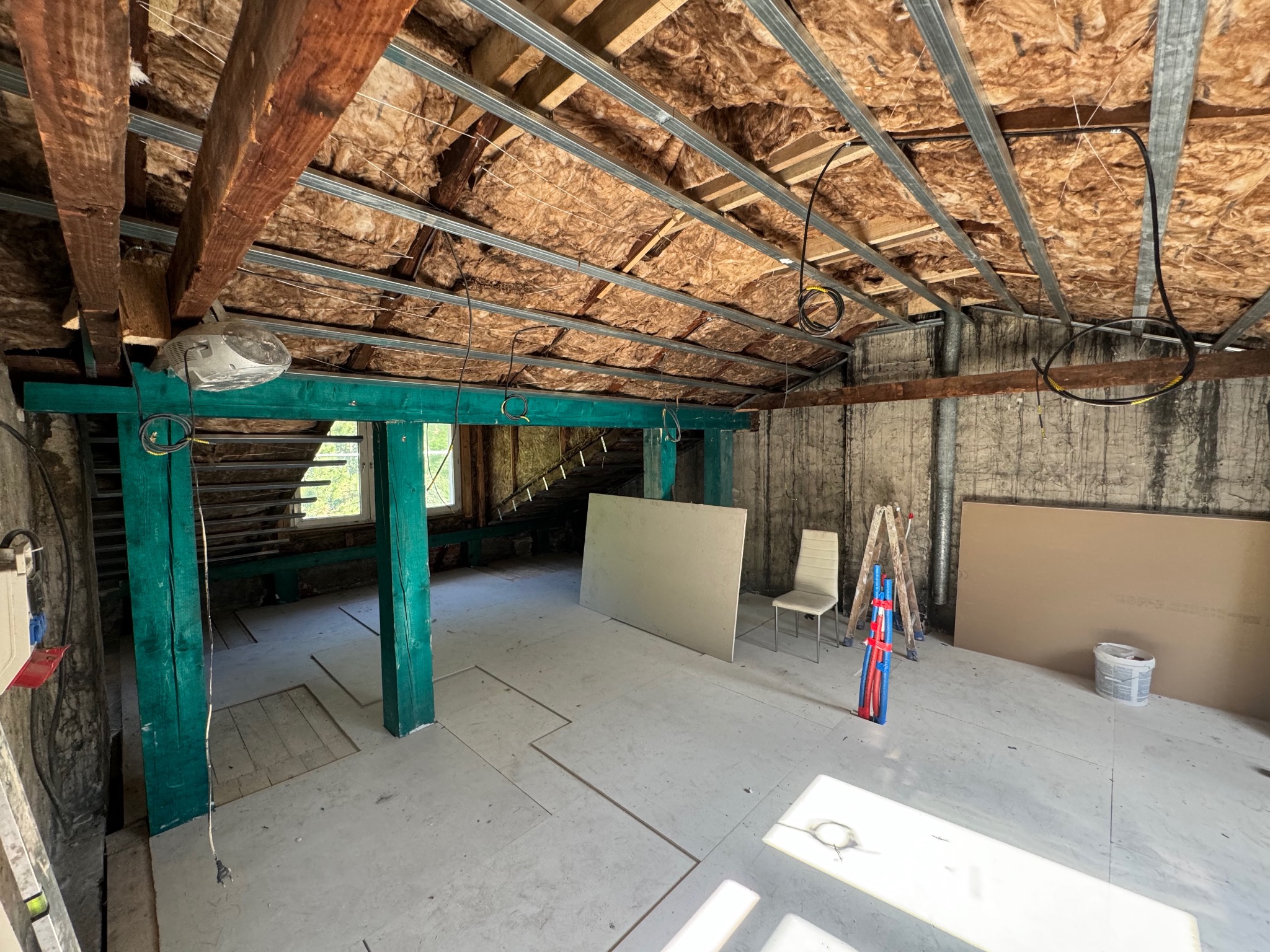 Poddasze w trakcie adaptacji: ocieplony dach z metalową konstrukcją, zielone drewniane słupy, płyty kartonowo-gipsowe oparte o ścianę, białe krzesło i drabina. Widoczne instalacje elektryczne...