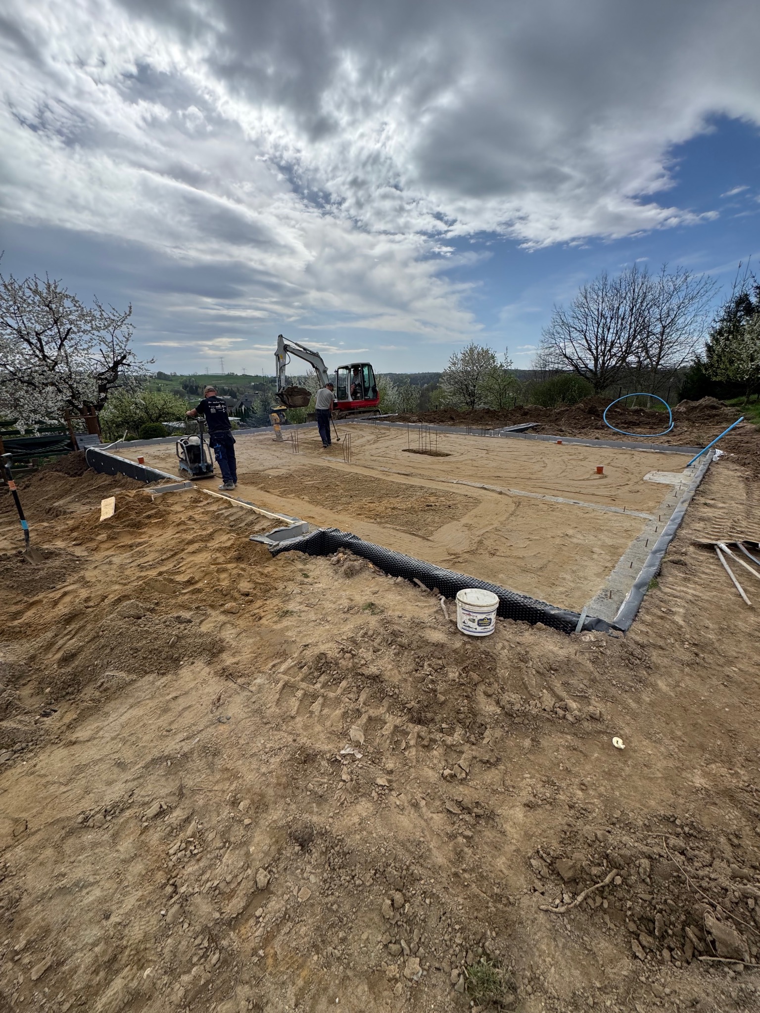 Przygotowanie terenu pod fundamenty domu jednorodzinnego: pracownicy zagęszczają grunt ubijarką, w tle widoczna koparka, na pierwszym planie wiadro z zaprawą.