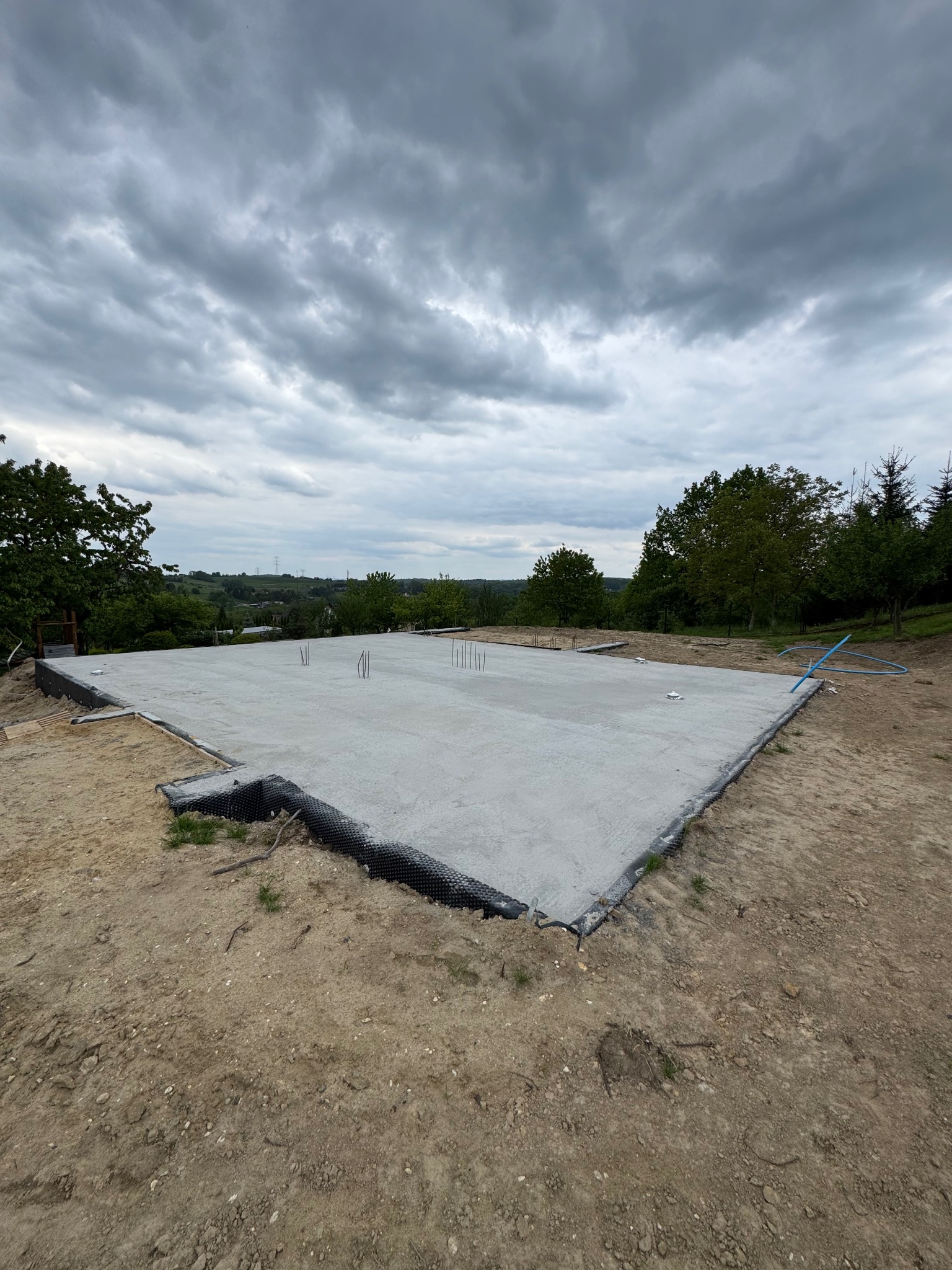 Wylana płyta fundamentowa na przygotowanym terenie, widoczne wystające pręty zbrojeniowe i hydroizolacja. Tło stanowią drzewa i pochmurne niebo.