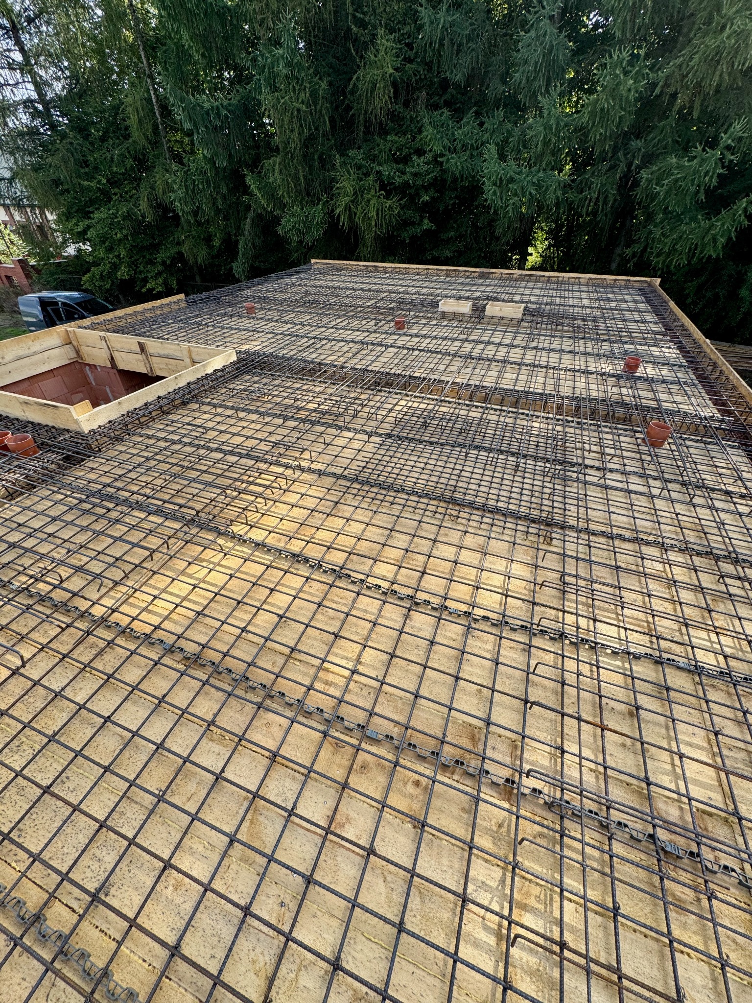 Zbrojenie stropu na deskowaniu z widocznymi elementami instalacji. W tle zieleń drzew. Kąt ujęcia z góry, ukazujący siatkę zbrojeniową i przygotowanie do betonowania.