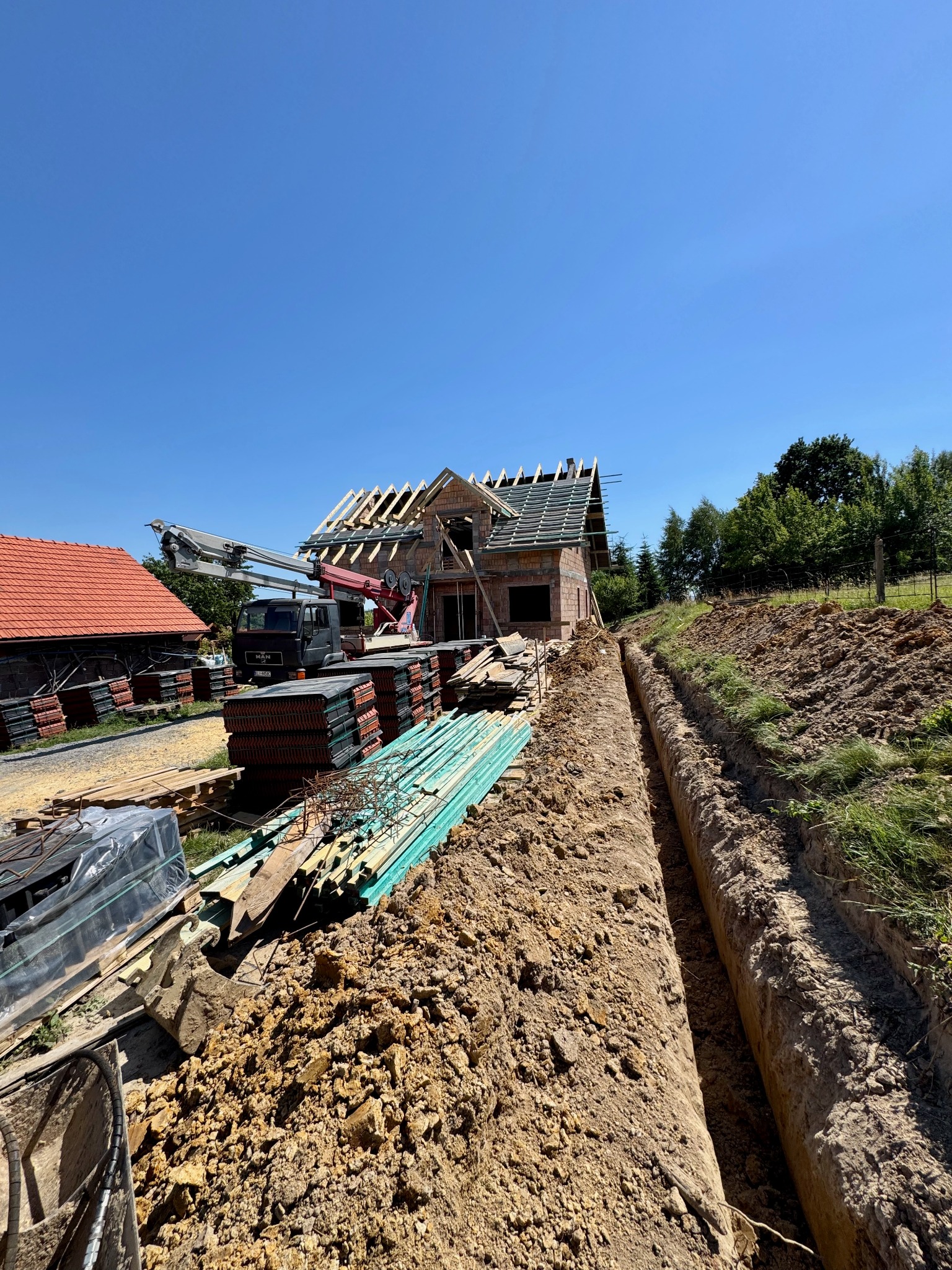 Budowa domu jednorodzinnego w trakcie realizacji: widoczny stan surowy otwarty, dach w budowie, wykop pod fundamenty, materiały budowlane składowane na placu budowy.