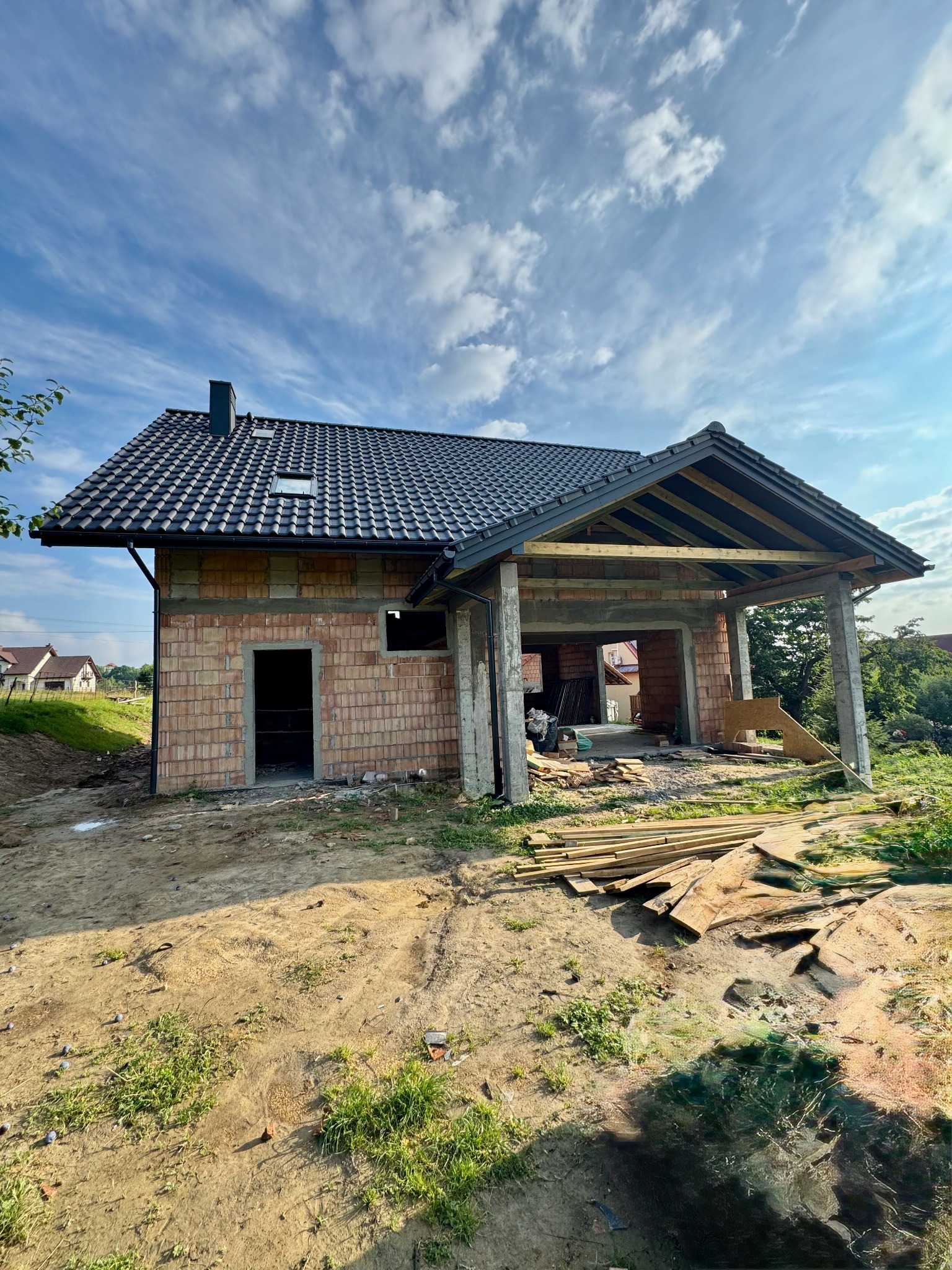 Budowa domu jednorodzinnego w stanie surowym otwartym z widocznym zadaszeniem nad miejscem parkingowym. Cegła ceramiczna, dachówka w kolorze grafitowym.