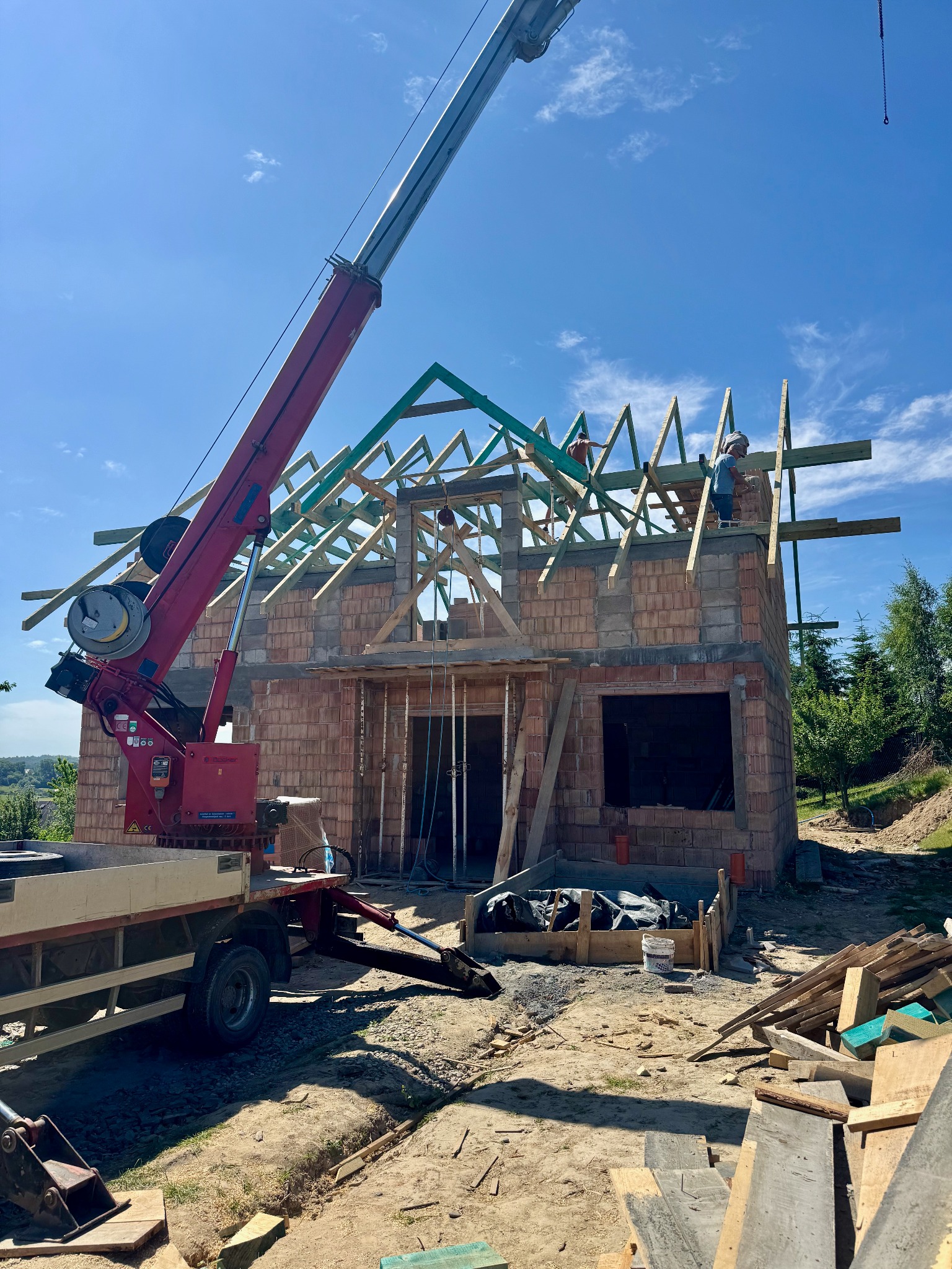 Budowa domu jednorodzinnego w trakcie wznoszenia dachu. Widoczny dźwig, murarz i cieśla pracujący na wysokości. Ceglany budynek w stanie surowym otwartym.