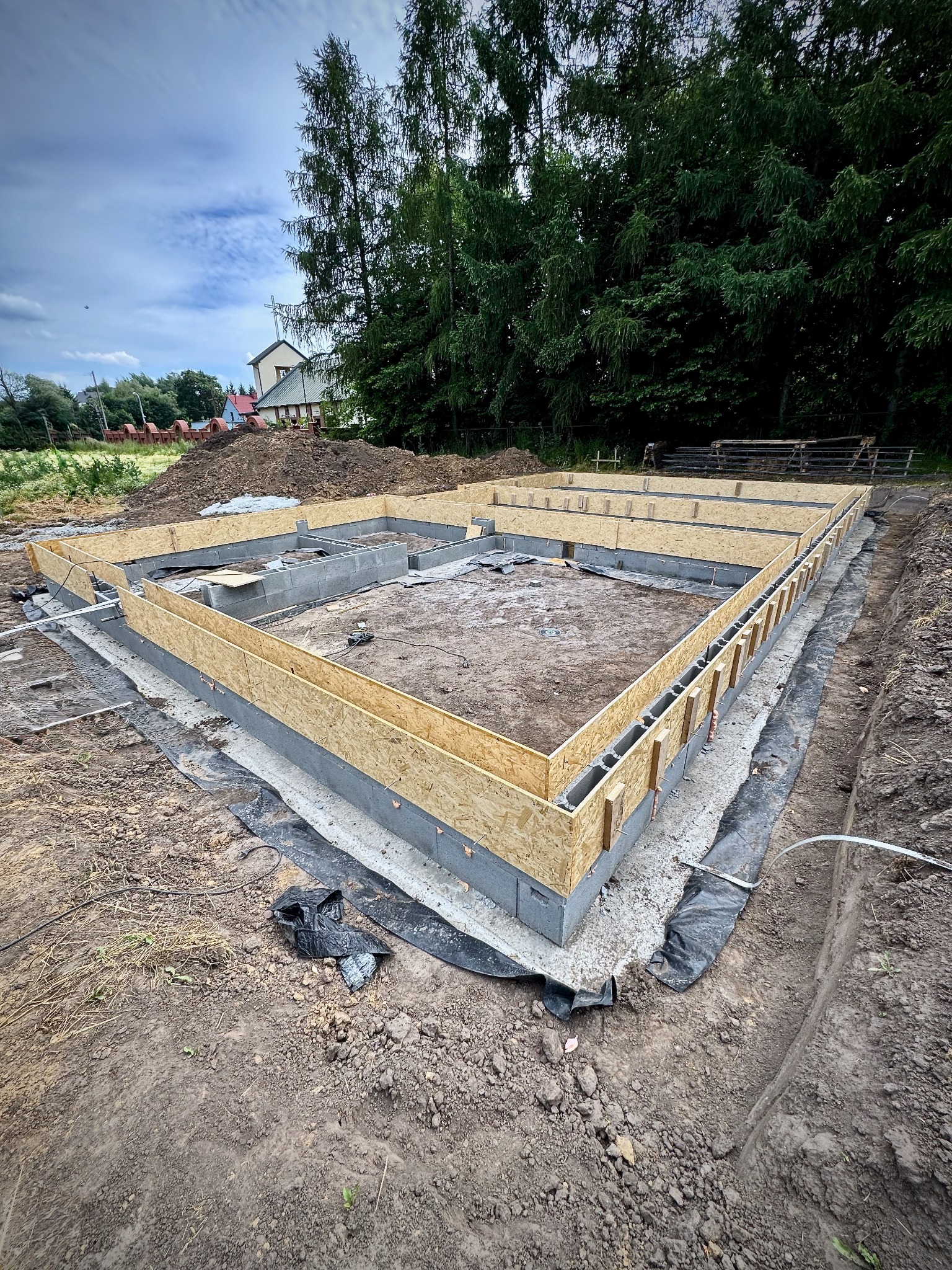 Fundamenty domu jednorodzinnego w trakcie budowy, z widoczną szalunką i bloczkami betonowymi. W tle zieleń drzew i fragment zabudowań.