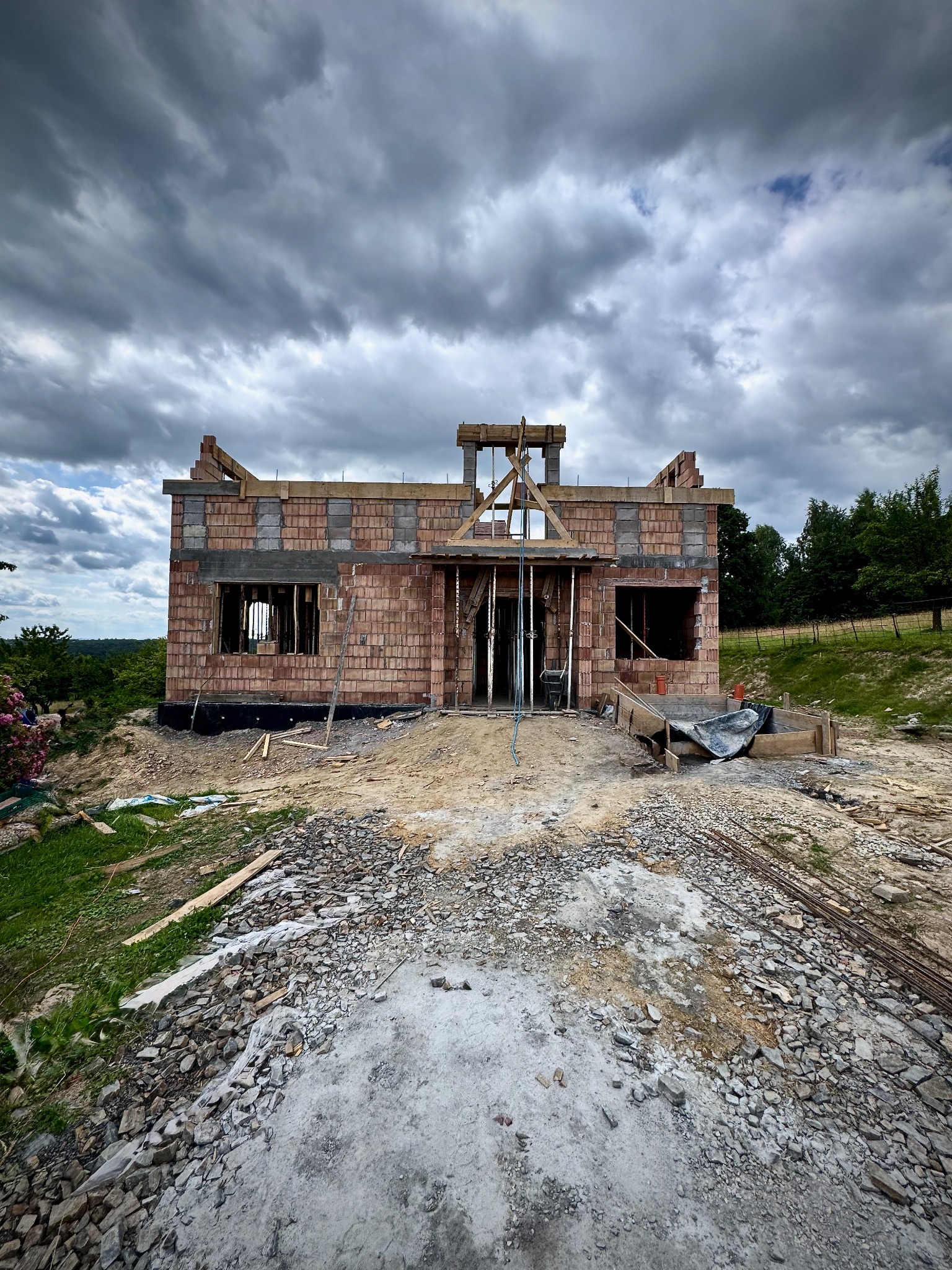 Surowy stan domu jednorodzinnego w trakcie budowy, widok z zewnątrz. Czerwona cegła, szare bloczki, drewniane elementy konstrukcyjne. Pochmurne niebo w tle.