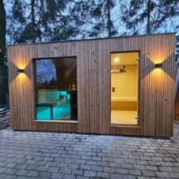 Nowoczesna, drewniana konstrukcja z oknem i otwartymi drzwiami, oświetlona zewnętrznymi lampami ściennymi, widoczna z zewnątrz na tle drzew i kostki brukowej.