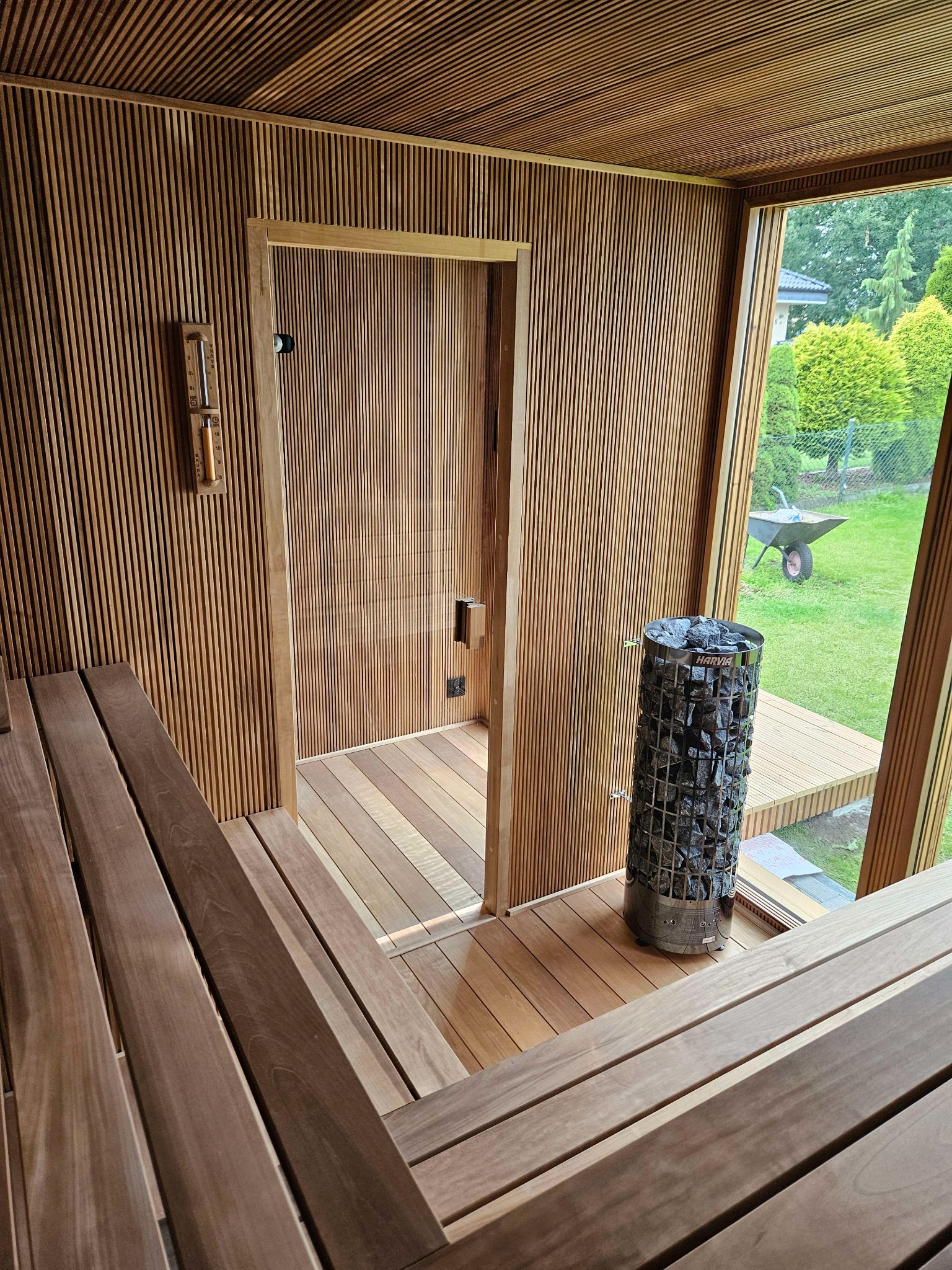 Wnętrze drewnianej kabiny z piecem na kamienie Harvia, widok na zewnątrz przez duże okno, gdzie widać trawnik i taczkę.