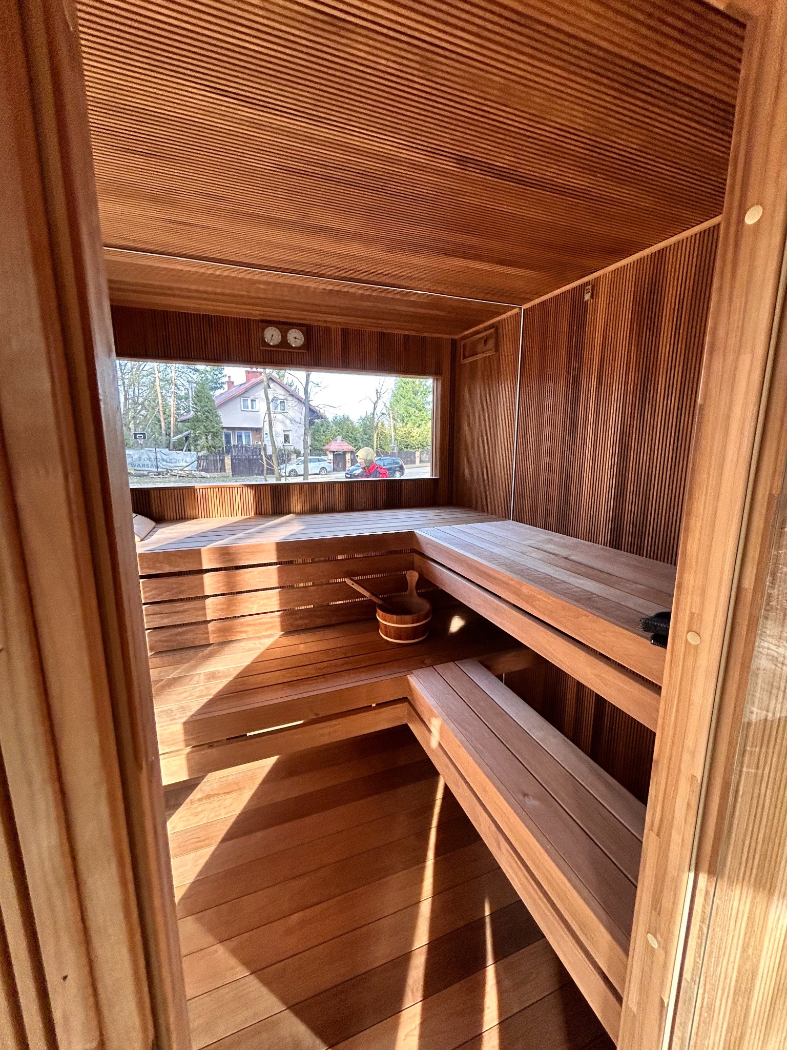 Wnętrze drewnianej kabiny z ławkami, wiadrem i oknem z widokiem na dom i ulicę.