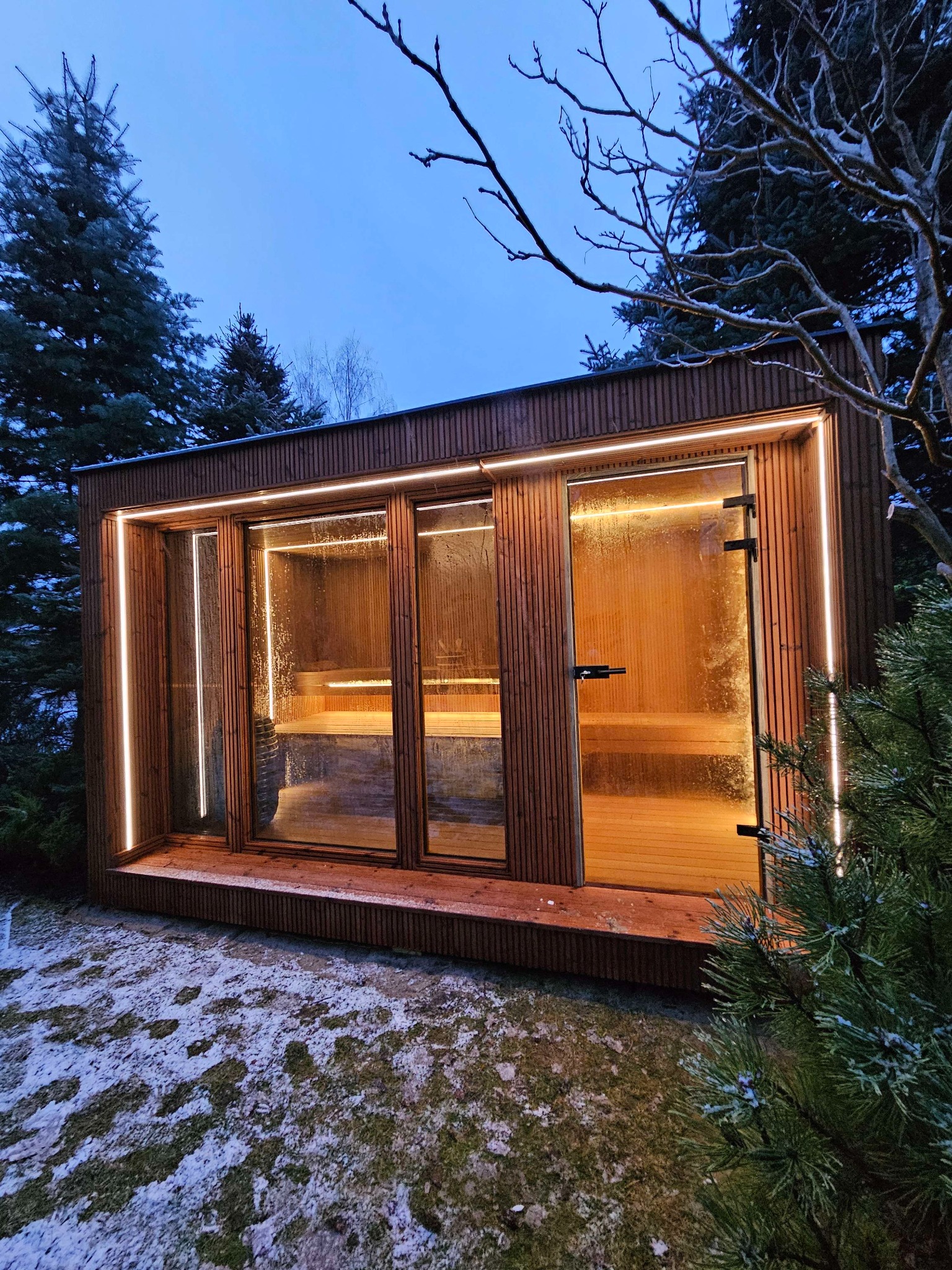 Nowoczesna drewniana kabina z przeszklonymi ścianami i oświetleniem LED, ustawiona na zewnątrz w zimowym otoczeniu z częściowo ośnieżonym trawnikiem i drzewami iglastymi.