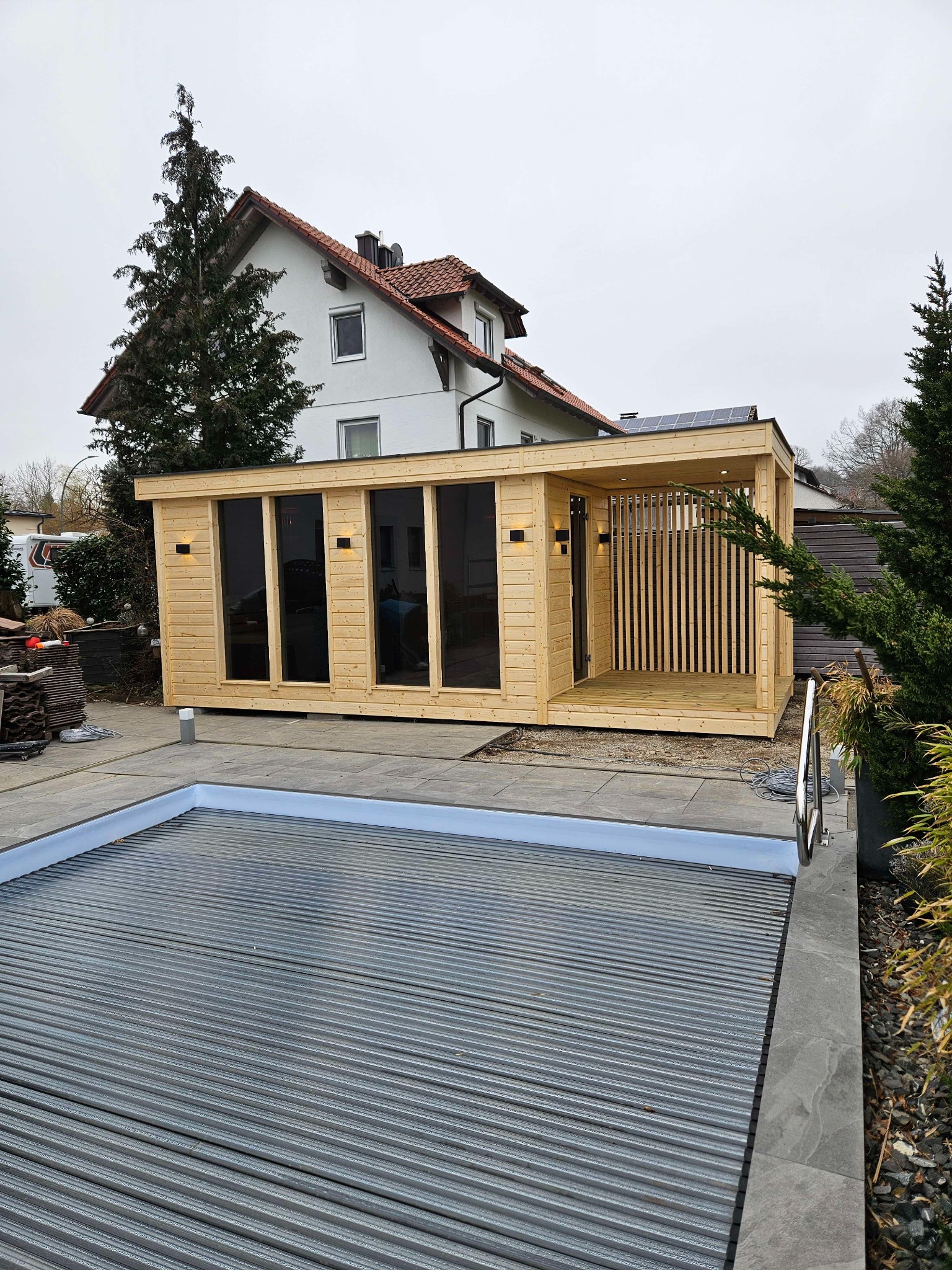 Nowoczesna, drewniana sauna ogrodowa z przeszklonymi ścianami i zadaszonym tarasem, obok zakrytego basenu na tle domu jednorodzinnego.