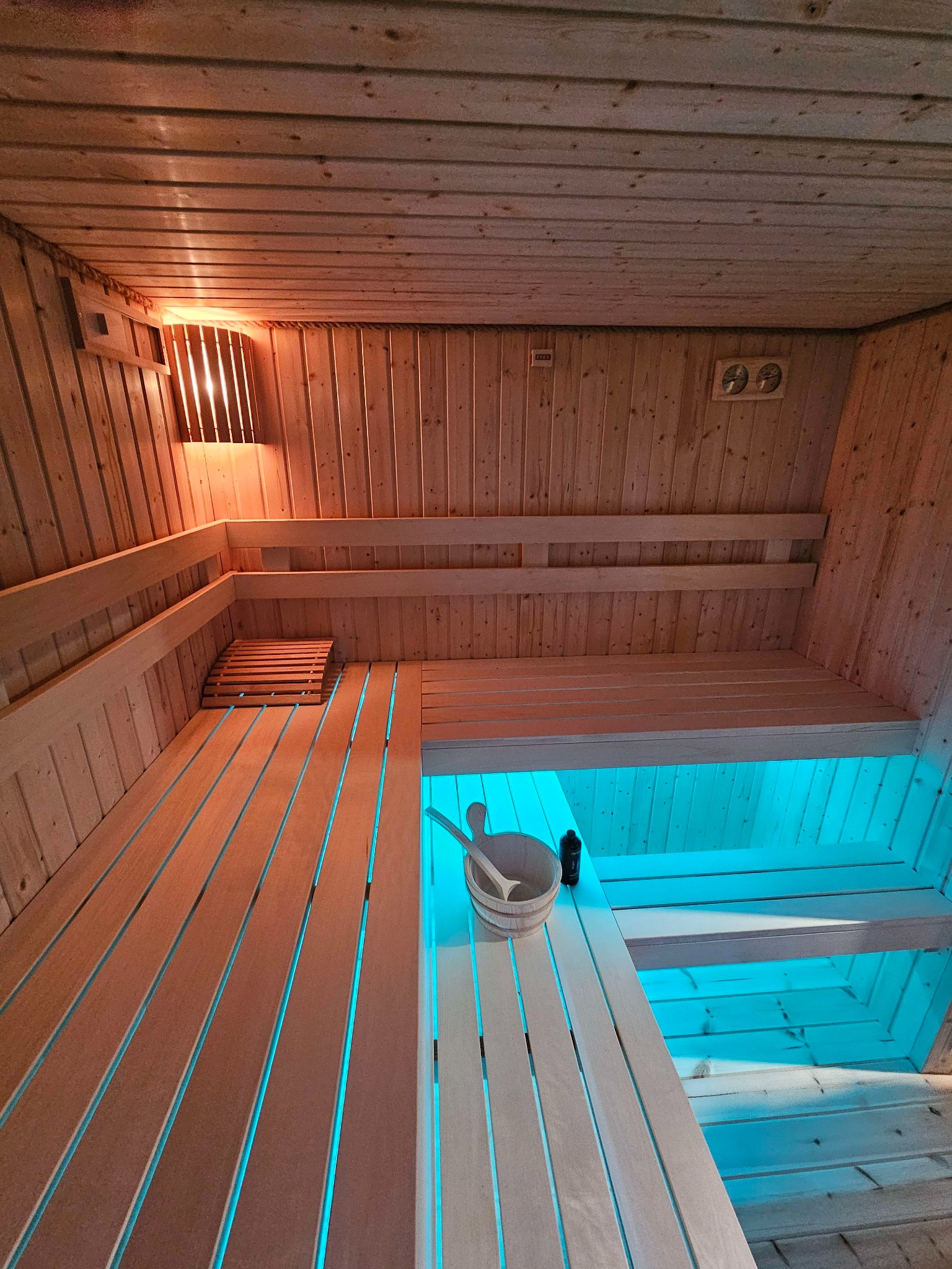 Wnętrze drewnianej kabiny z oświetleniem LED w kolorze turkusowym, widoczny drewniany kubeł z chochlą i butelka.