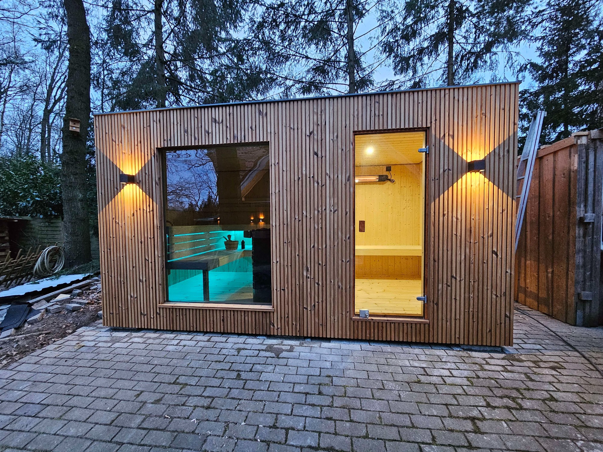 Nowoczesna, drewniana konstrukcja z oknem i otwartymi drzwiami, oświetlona zewnętrznymi lampami ściennymi, widoczna z zewnątrz na tle drzew i kostki brukowej.