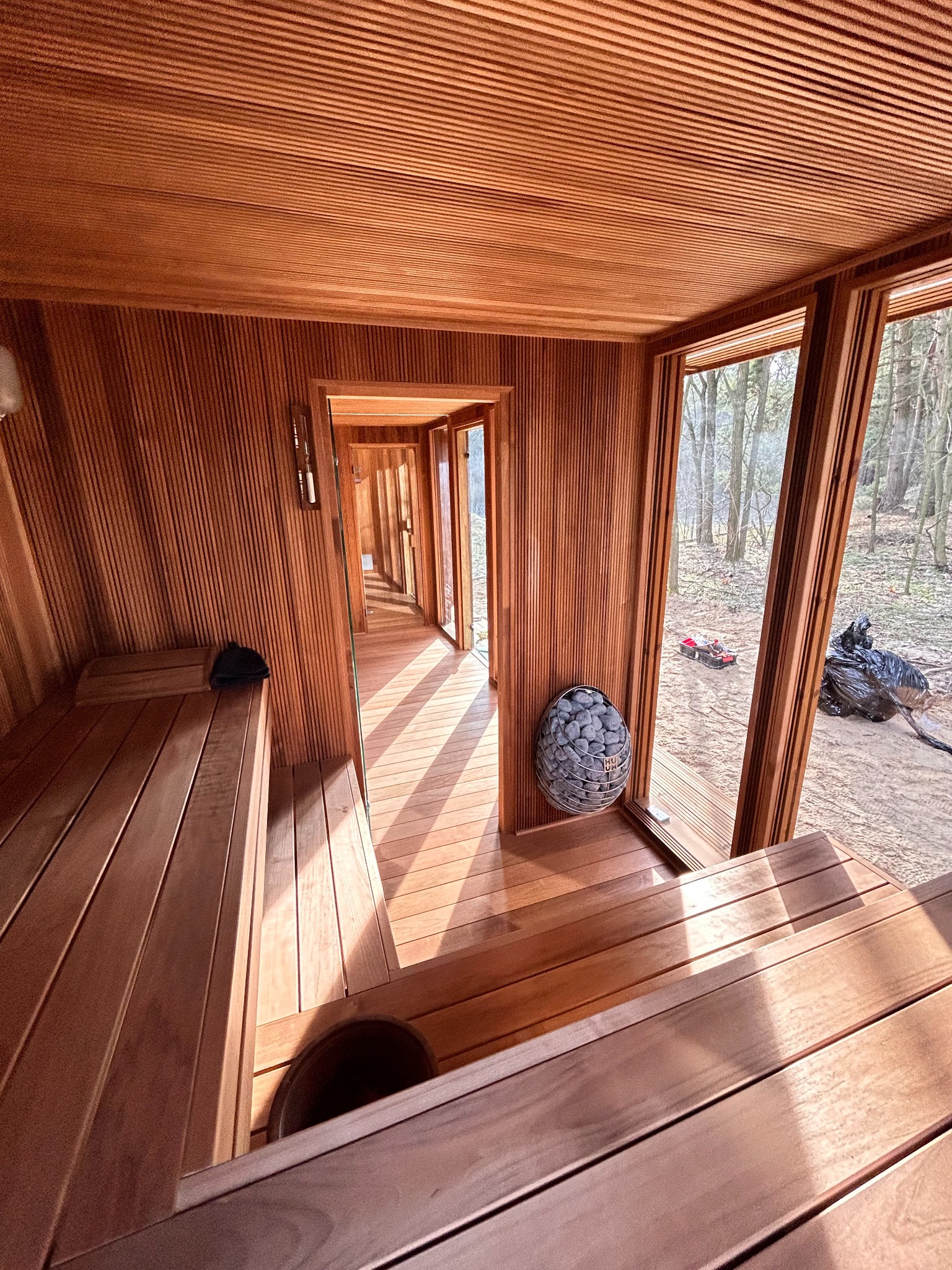 Wnętrze drewnianej kabiny z ławkami, widok na las przez okna, kosz z kamieniami do pieca.