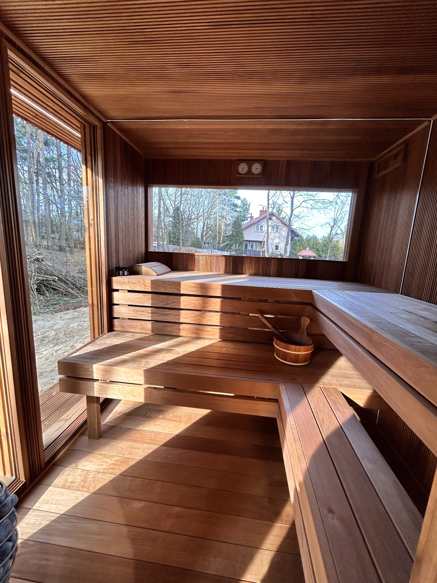 Wnętrze drewnianej kabiny z widokiem na zewnątrz przez okno, ukazujące drzewa i dom. Na ławkach stoi drewniane wiadro z chochlą, a na górnej półce leży poduszka i kilka małych butelek.