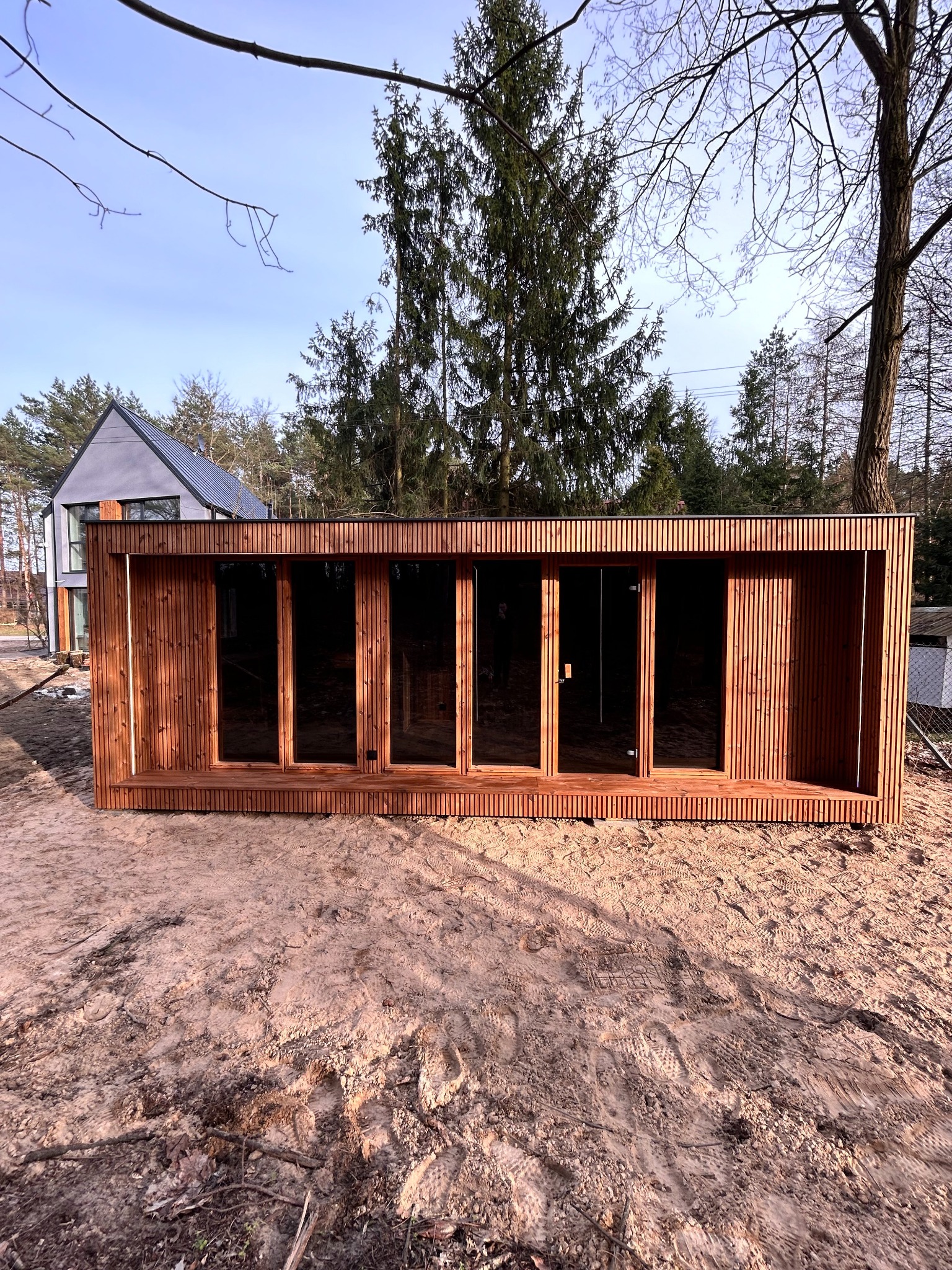 Drewniana konstrukcja z przeszklonymi drzwiami na piaszczystym terenie, nowoczesny dom w tle, widok z zewnątrz.