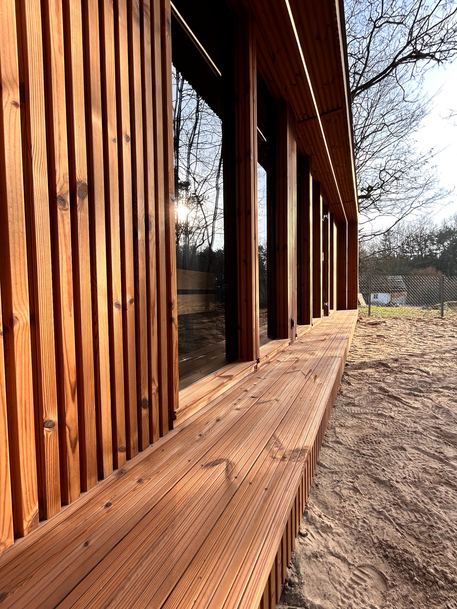 Nowoczesny drewniany budynek z lamelową fasadą i tarasem, widok z perspektywy ziemi, z widocznym słońcem przebijającym przez drzewa w tle.