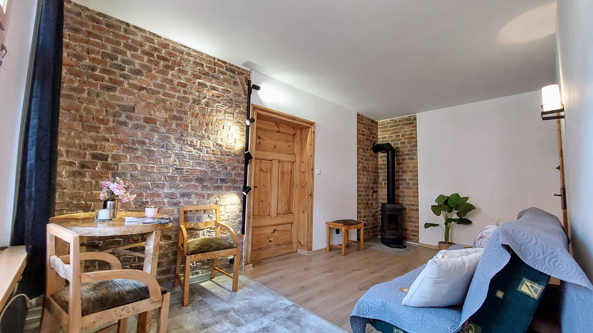 Rustykalne wnętrze: ceglana ściana, drewniany stół z kwiatami, piec typu koza. Stylizacja mieszkania po usłudze najmu, widok na salon z elementami dekoracyjnymi.