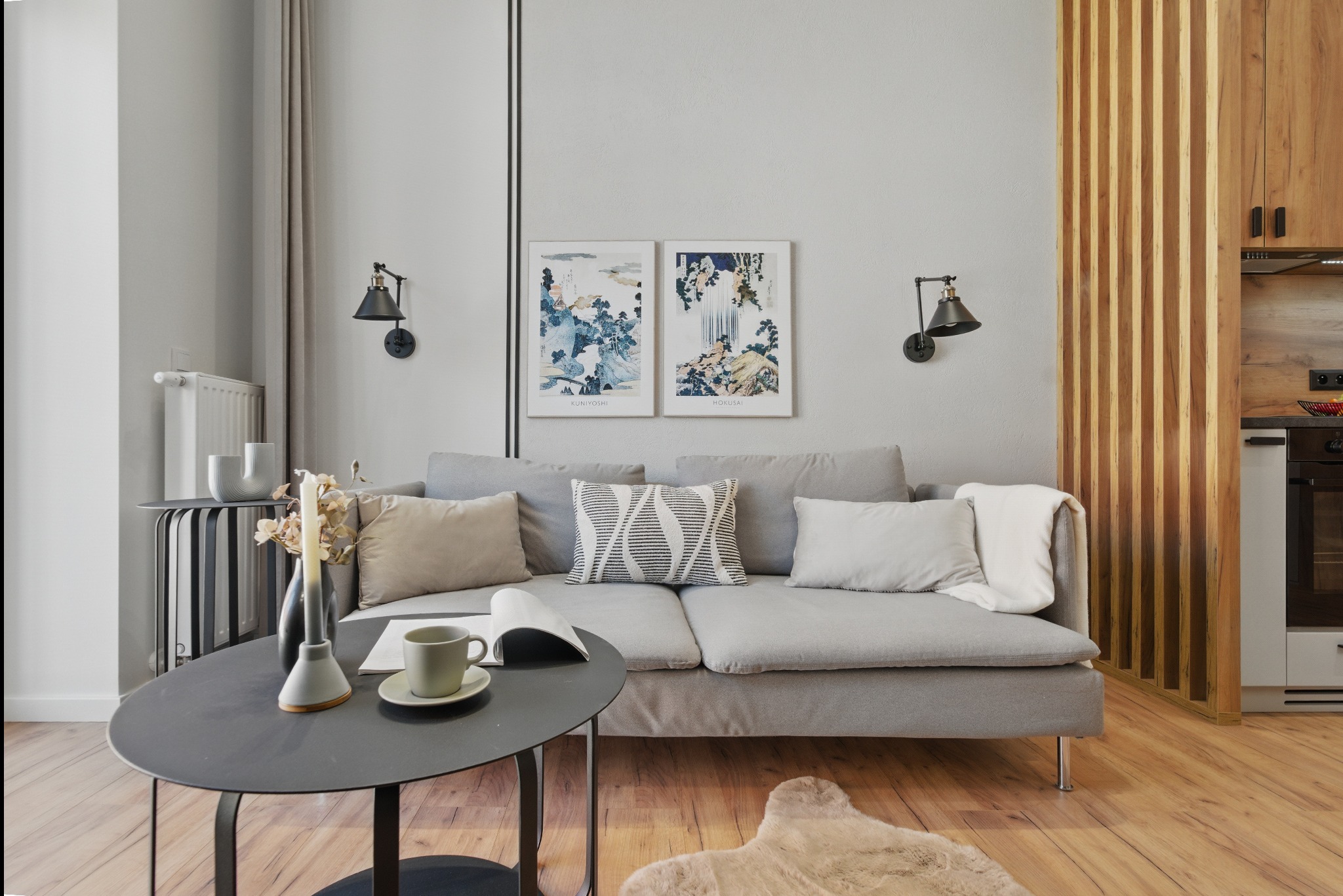 Jasne wnętrze mieszkania z szarą sofą, stolikiem kawowym i drewnianymi akcentami, ukazujące estetyczne wykończenie i aranżację, idealne do najmu.