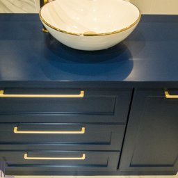 Kuchnie na wymiar Warszawa DMKUCHNIE - Niebieska szafka łazienkowa z trzema szufladami i umywalką nablatową z złoconym rantem, widoczne złote uchwyty i fragment marmurowej ściany