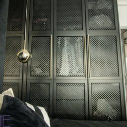 Kuchnie na wymiar Warszawa DMKUCHNIE - Czarna szafa wnękowa z perforowanymi drzwiami i złotymi uchwytami, widoczna z perspektywy łóżka, z lampą wiszącą w kształcie kuli i fragmentem zasłony.