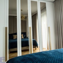 Kuchnie na wymiar Warszawa DMKUCHNIE - Biała szafa wnękowa z lustrzanymi drzwiami, odbijająca wnętrze sypialni z niebieskim łóżkiem i złotym żyrandolem.