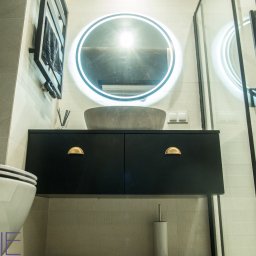 Kuchnie na wymiar Warszawa DMKUCHNIE - Nowoczesna łazienka z umywalką w kształcie misy, podświetlanym okrągłym lustrem, czarną szafką pod umywalką z półokrągłymi złotymi uchwytami i kabiną prysznicową z czarnymi ramami.