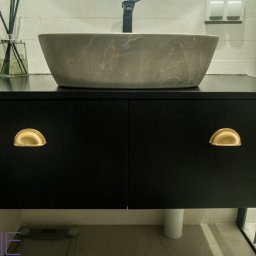 Kuchnie na wymiar Warszawa DMKUCHNIE - Szara umywalka w kształcie misy na czarnej szafce z dwoma drzwiczkami i złotymi uchwytami w łazience z jasnymi płytkami.