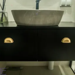 Szara umywalka w kształcie misy na czarnej szafce z dwoma drzwiczkami i złotymi uchwytami w łazience z jasnymi płytkami.