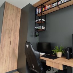 Kuchnie na wymiar Warszawa DMKUCHNIE - Pokój gracza z biurkiem z drewnianym blatem, krzesłem gamingowym, dwoma monitorami, półkami z książkami i szafą z drewnianymi frontami, utrzymany w szarej kolorystyce.