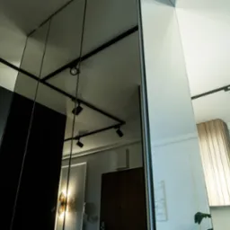 Częściowe ujęcie nowoczesnej zabudowy z czarnymi frontami, odbijającej się w lustrze, z widocznym logo firmy na dole po lewej stronie i oświetleniem szynowym na suficie.