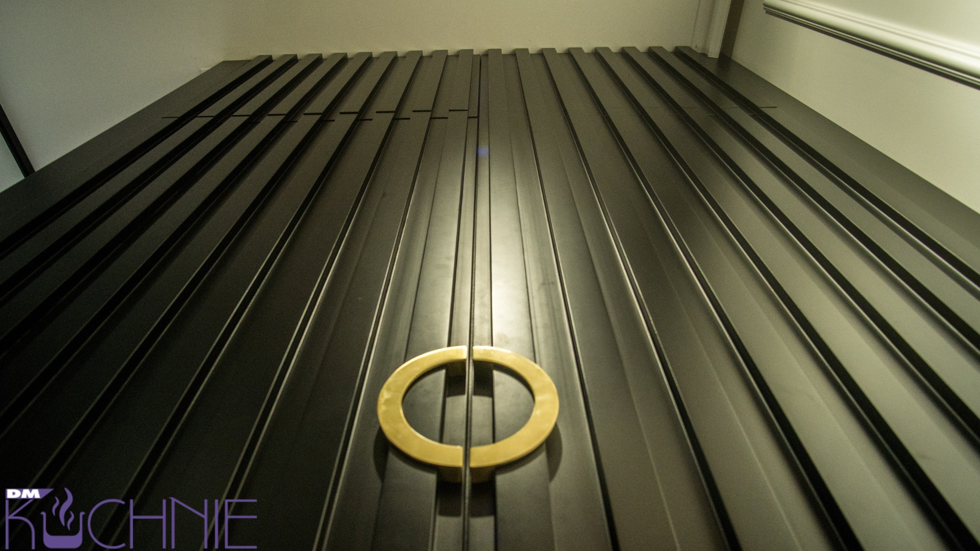 Ciemna, pionowo żłobkowana szafa z okrągłym, złotym uchwytem, widziana z dołu, z logo firmy w lewym dolnym rogu.