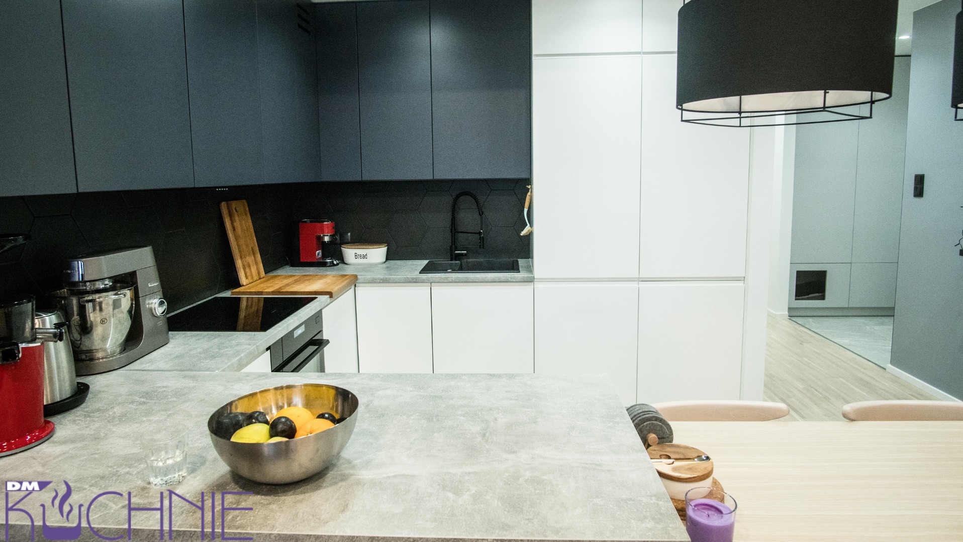 Nowoczesna kuchnia z szarymi górnymi szafkami i białymi dolnymi, czarnym panelem z heksagonalnych płytek nad blatem, wyspą z misą owoców i widokiem na jadalnię z lampami.